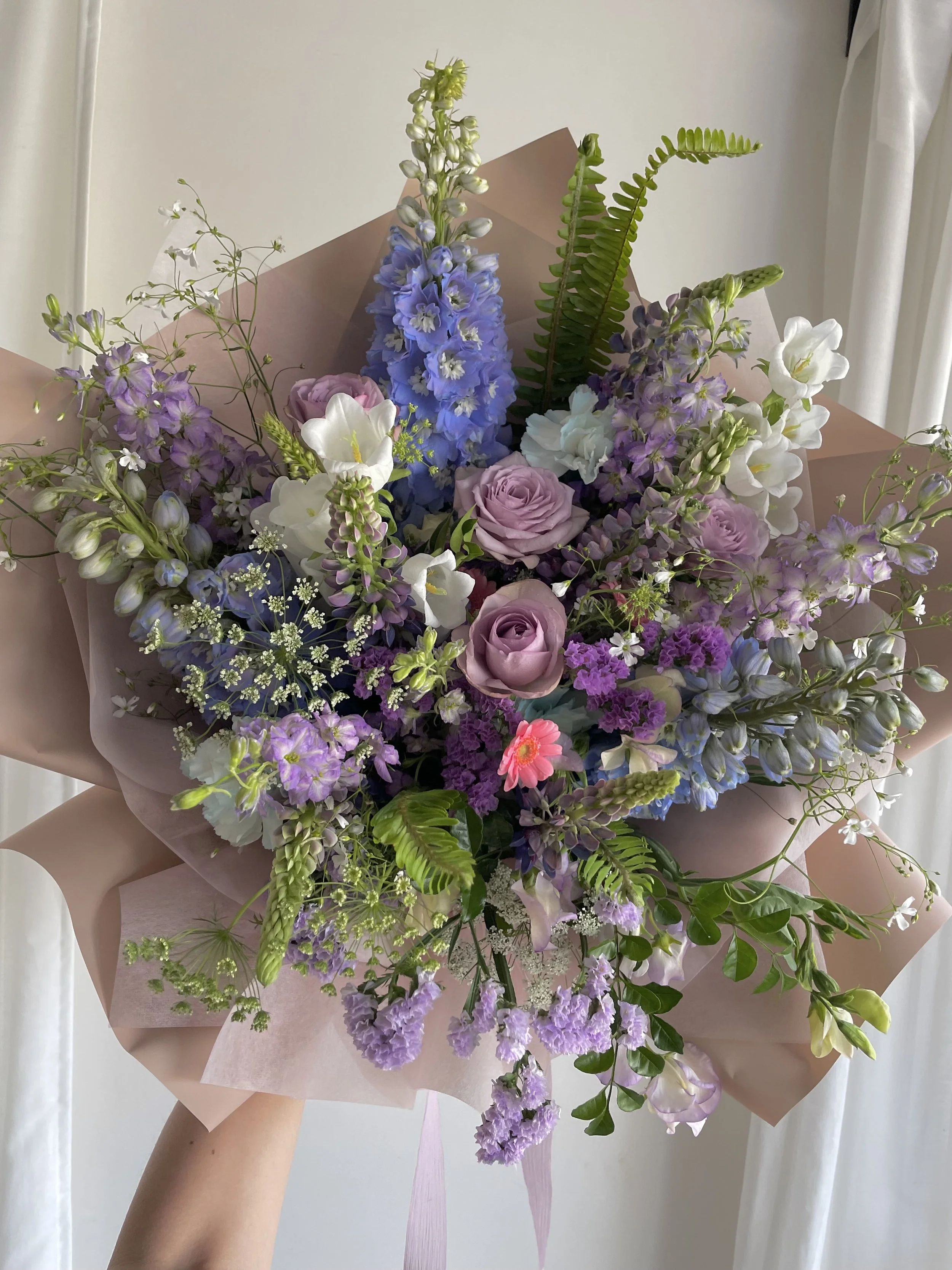 A colorful bouquet of various flowers including purple roses, lavender, blue delphiniums, and white lilies wrapped in pink paper.