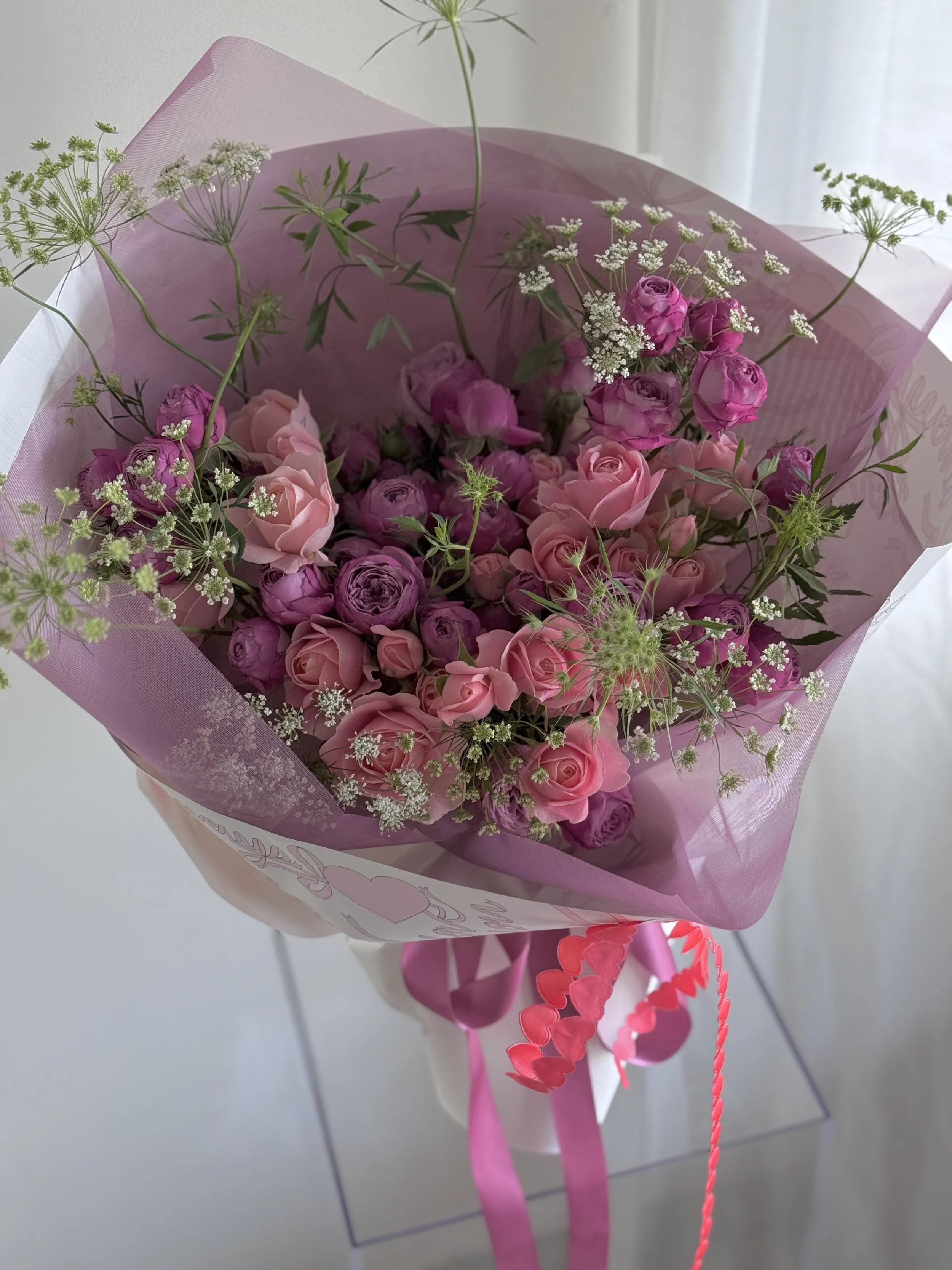 A bouquet of pink and purple roses with white baby's breath flowers and green foliage, wrapped in pink and white paper with pink ribbons.