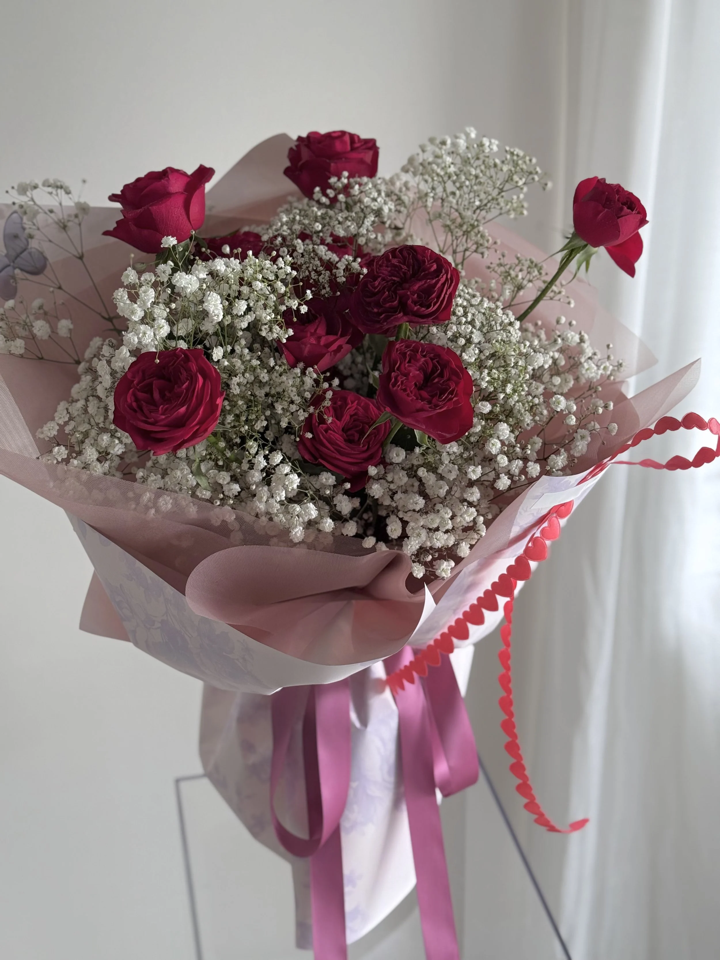 Bouquet of pink roses and white baby's breath flowers, wrapped in pink and white paper with pink ribbons, and a red heart-shaped curly ribbon.