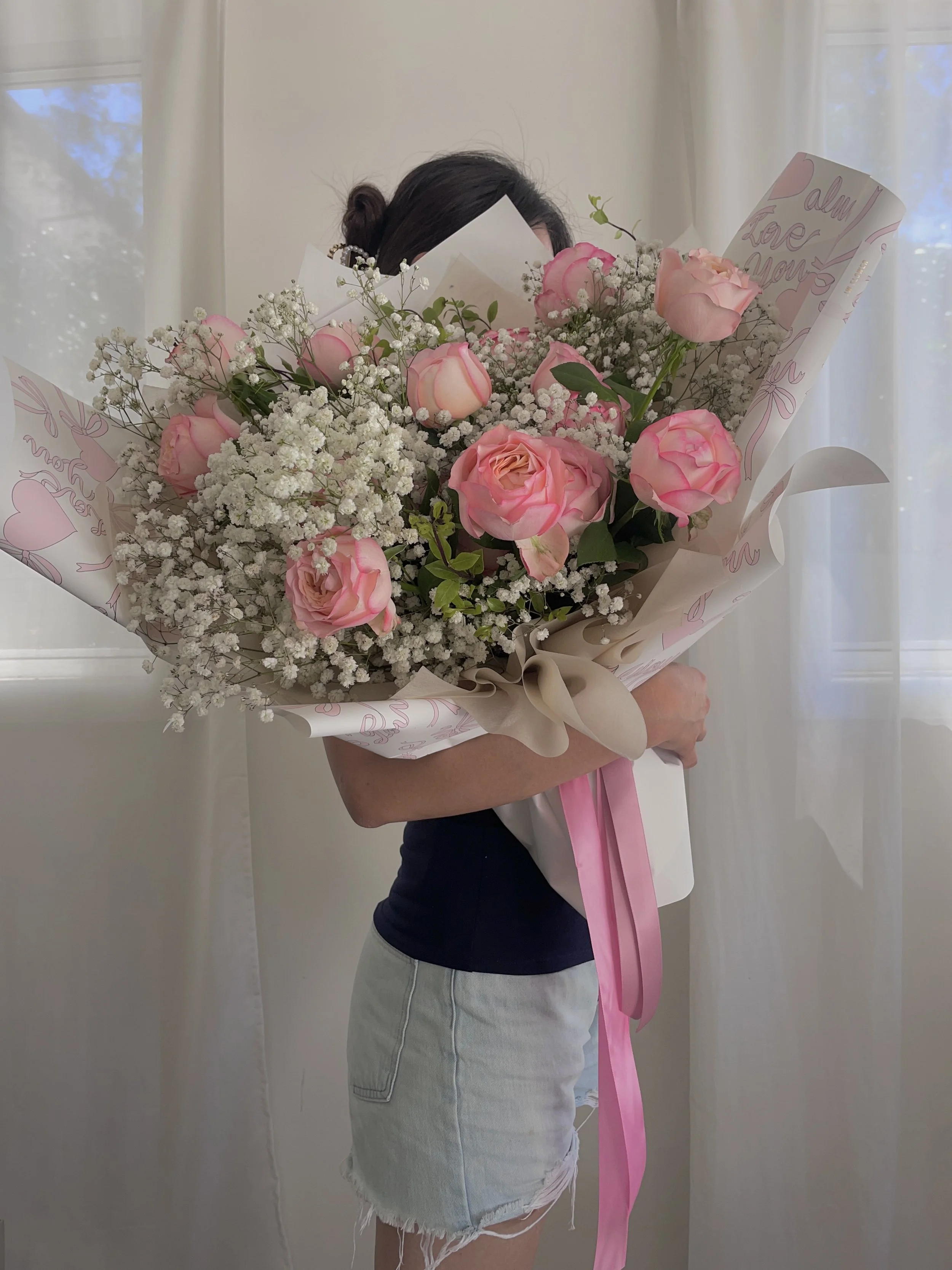 A woman holding a large bouquet of pink roses, white baby's breath, and greenery, wrapped in decorative pink and white paper with a pink ribbon.