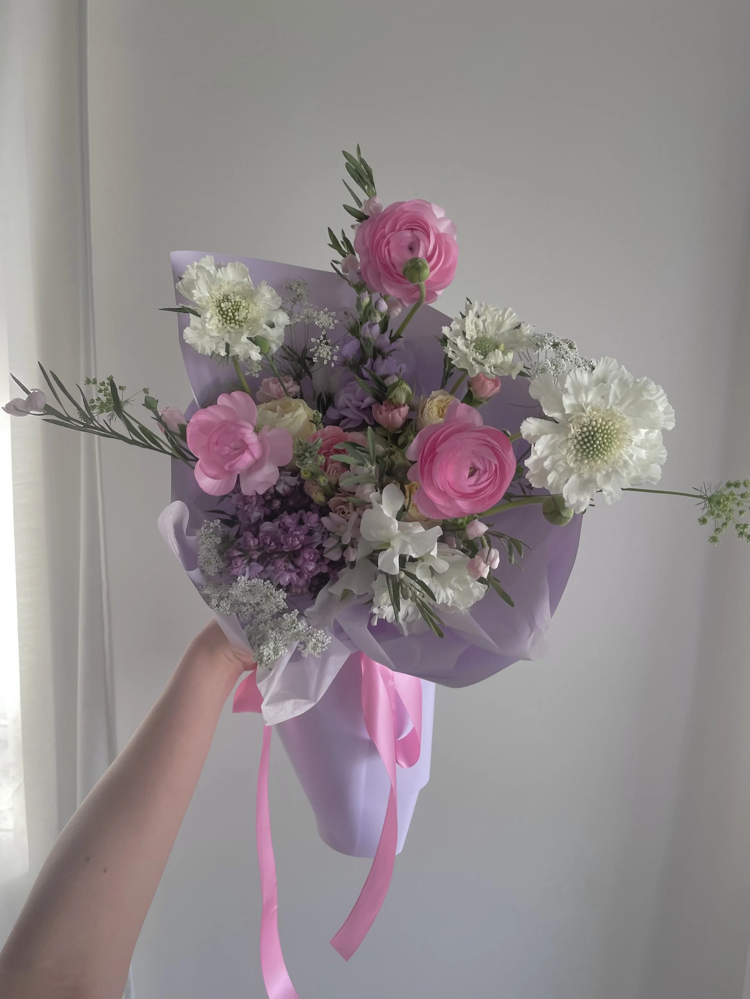 Hand holding a bouquet of pink, white, and purple flowers wrapped in purple paper with pink ribbon.