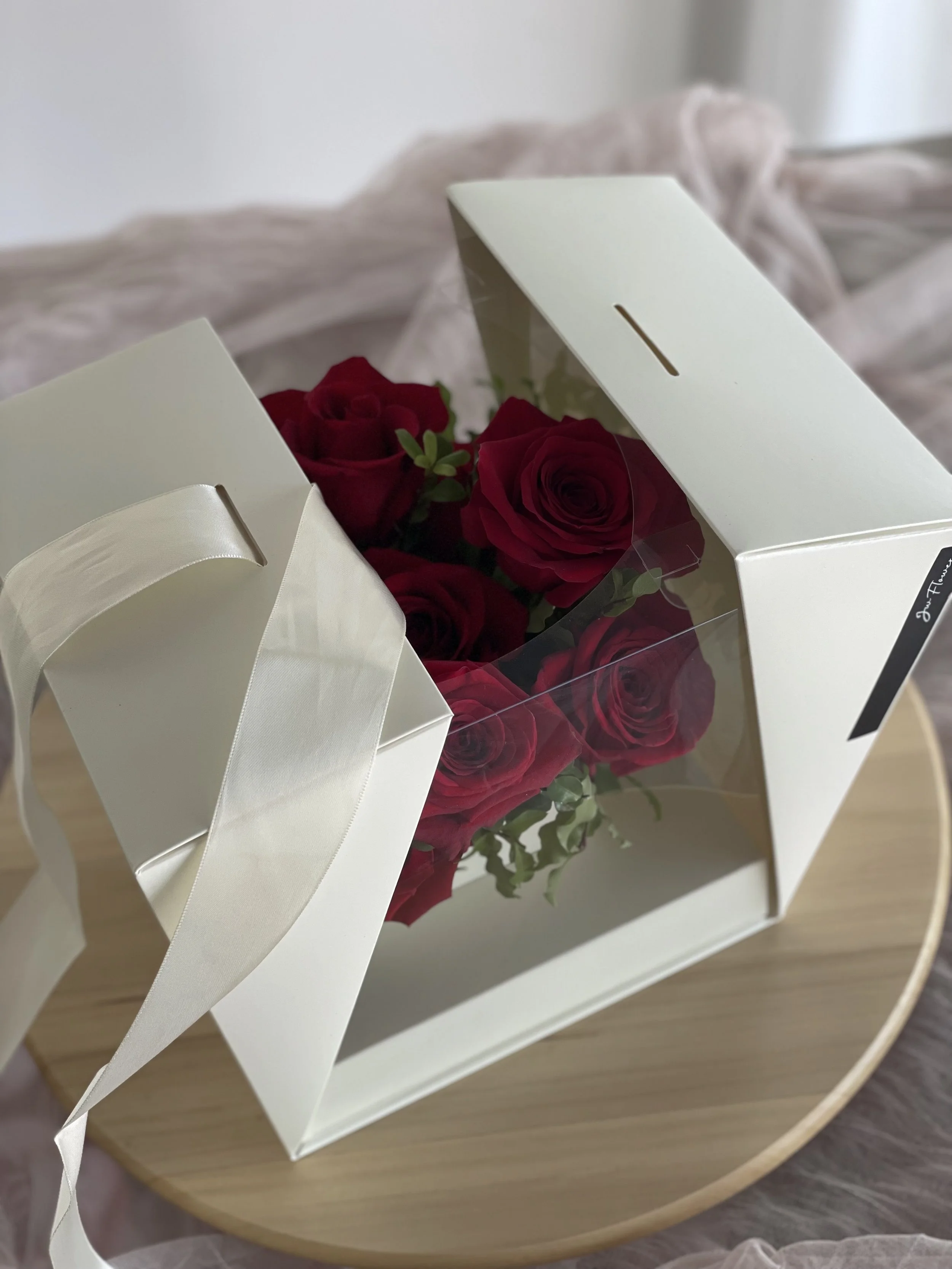 A bouquet of red roses in a clear plastic box with a white lid, decorated with a white ribbon, placed on a round wooden table.