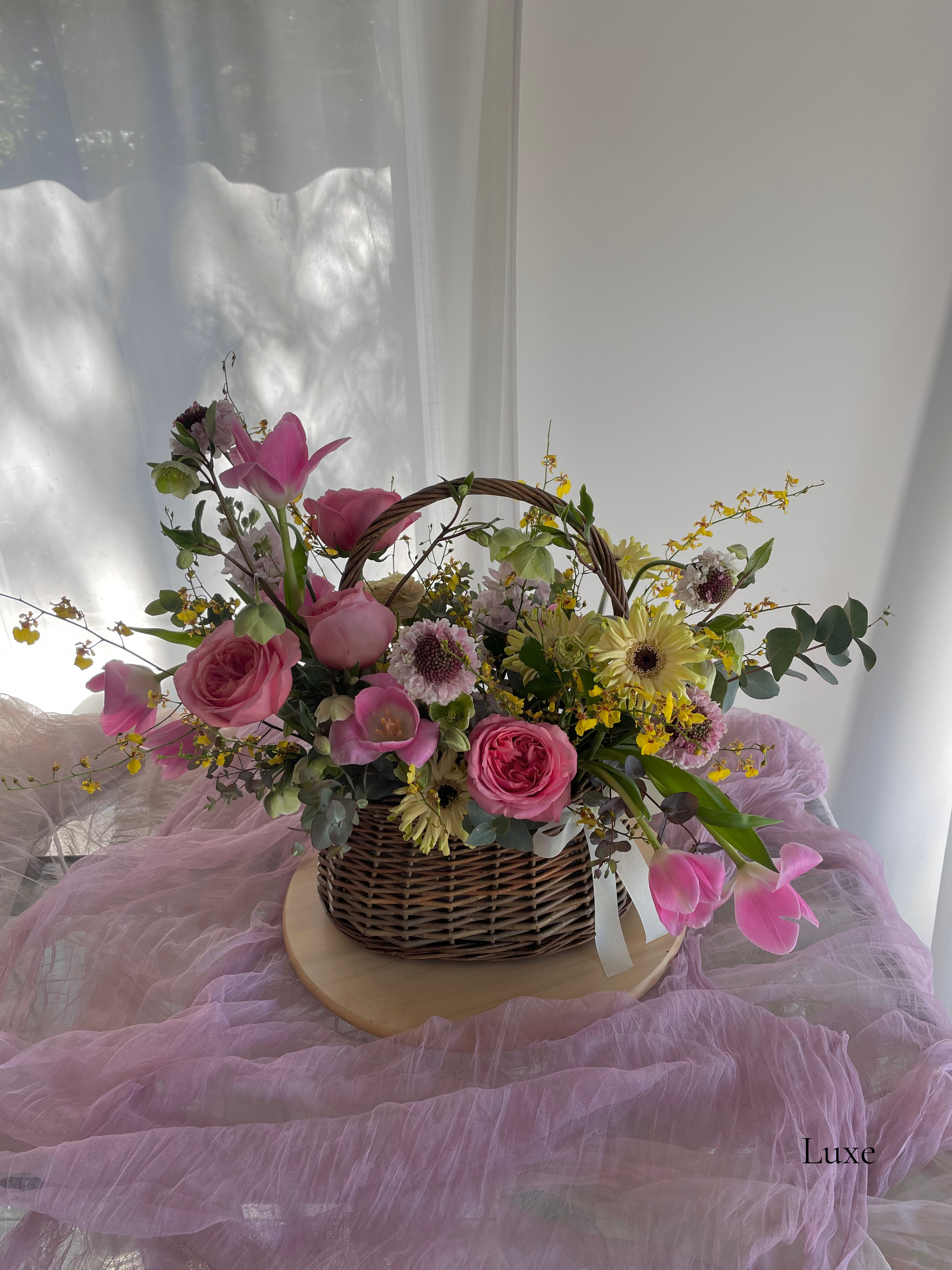 A floral arrangement in a wicker basket with pink roses, tulips, and other flowers on a round wooden platform, draped with pink fabric, set near a bright window.