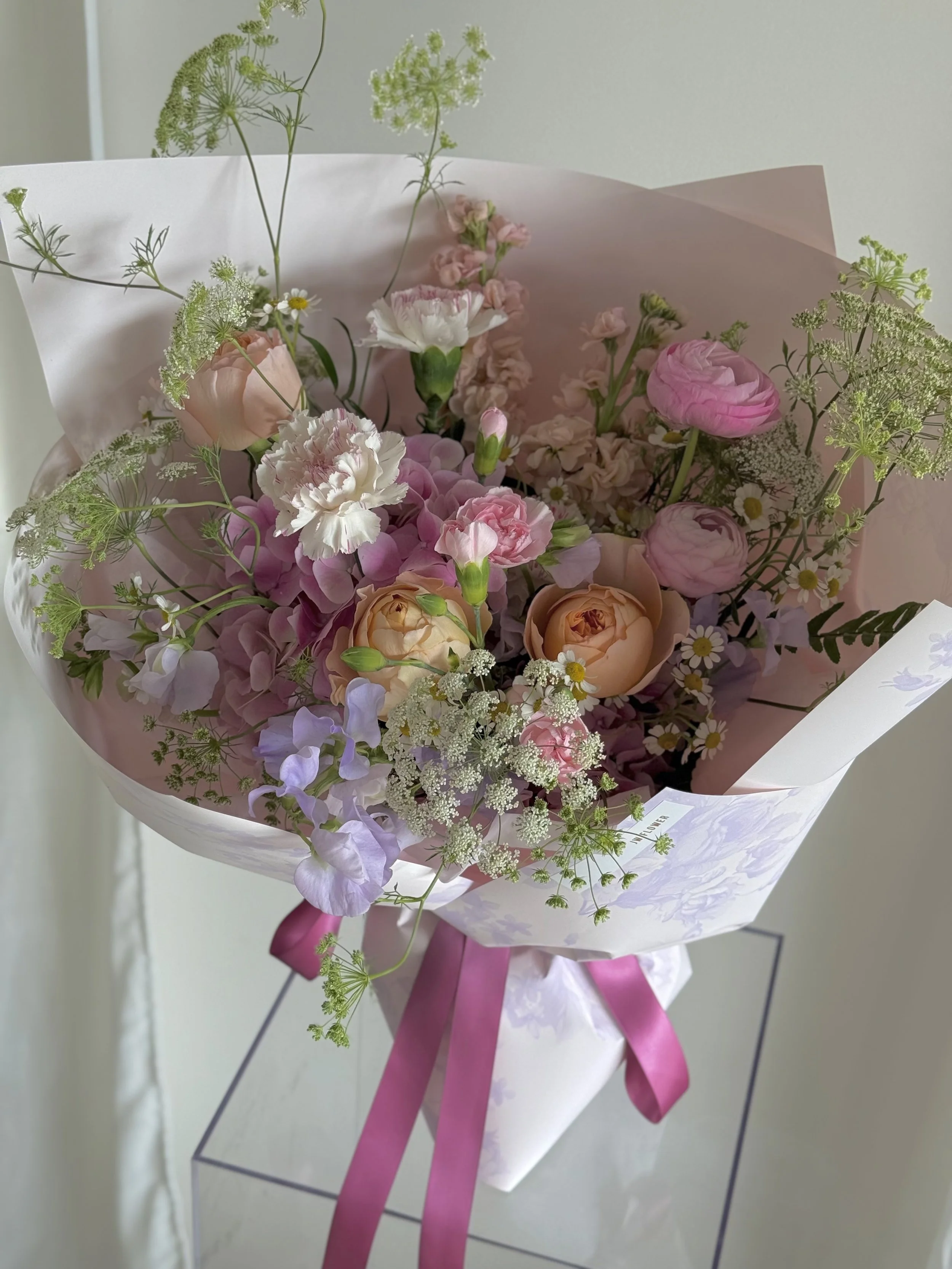 A bouquet of pastel-colored flowers wrapped in pink paper with a purple ribbon, placed on a glass stand.