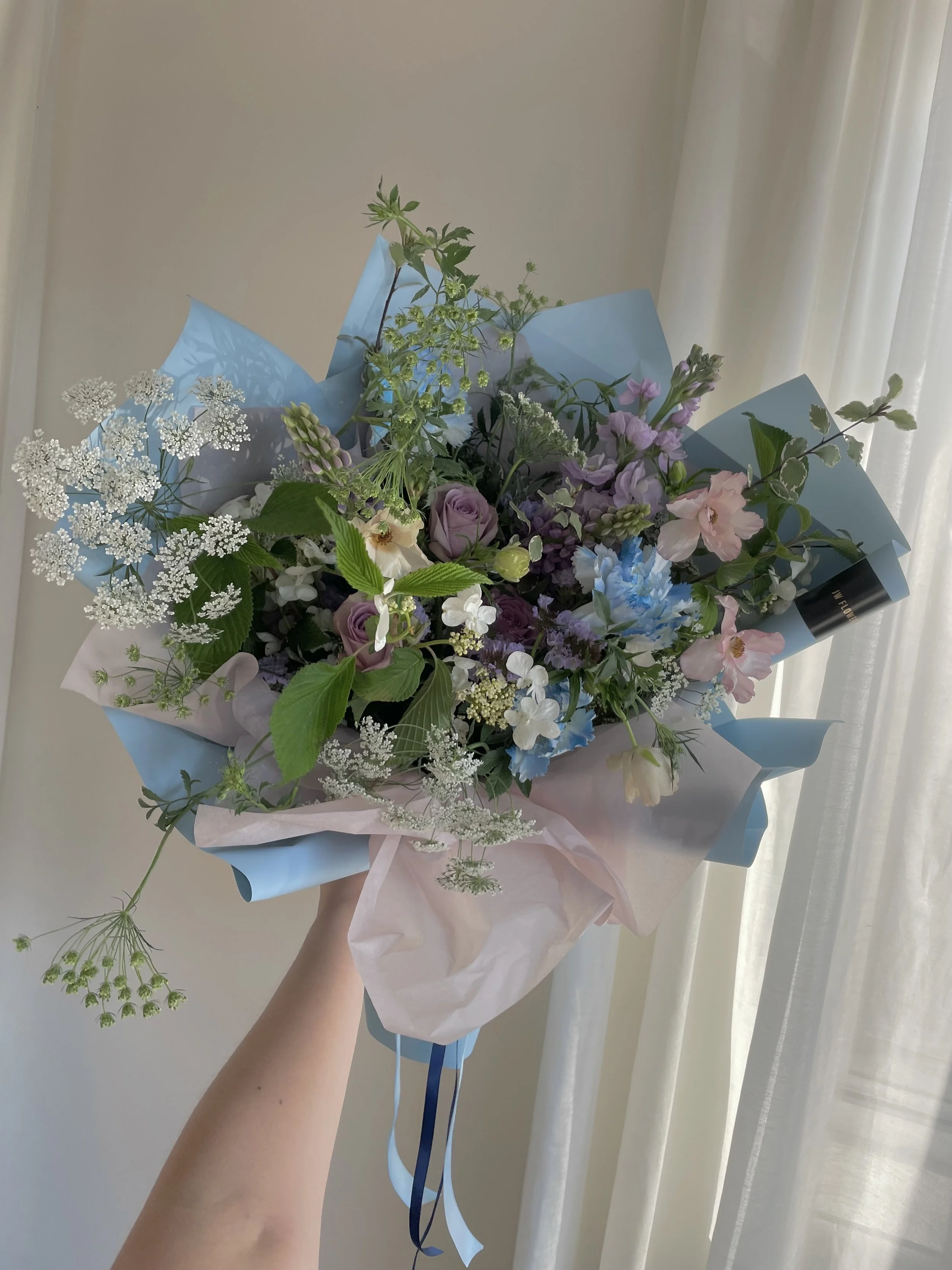 Hand holding a bouquet of mixed flowers wrapped in pastel pink and blue paper, with various purple, white, pink, and blue flowers, and green foliage, against a neutral background with light curtains.
