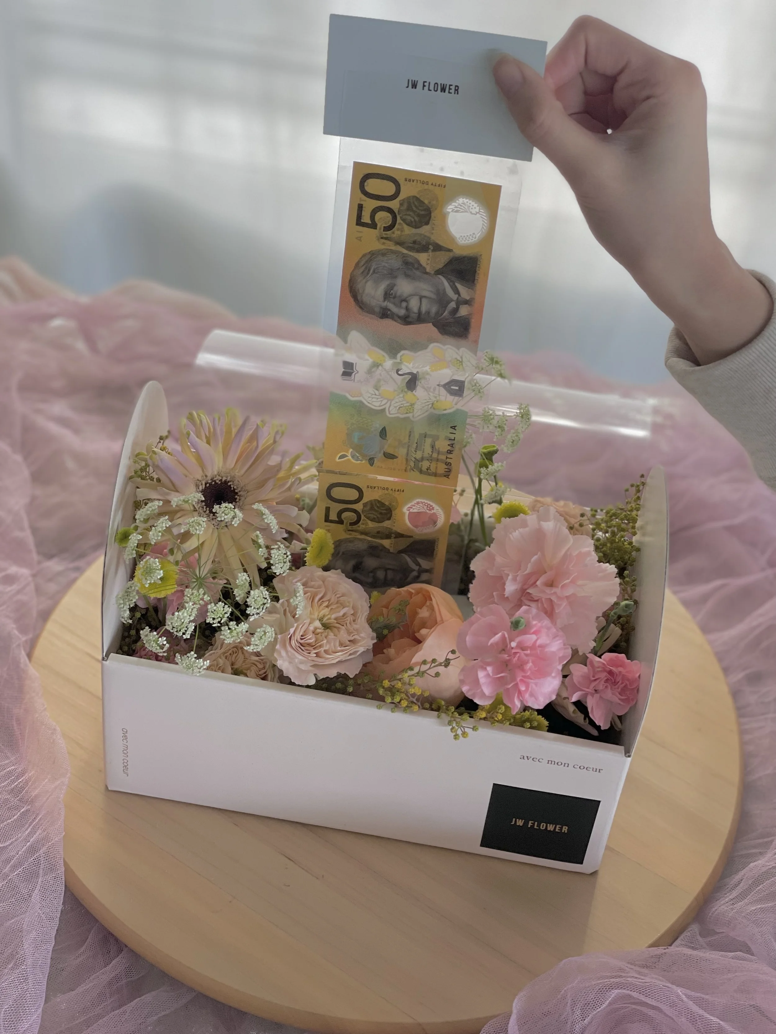 A white flower box with pink and purple flowers, a 50-dollar Australian banknote, and a person holding a small card that says "JW FLOWER." The box is on a round wooden surface, draped with pink tulle or fabric.