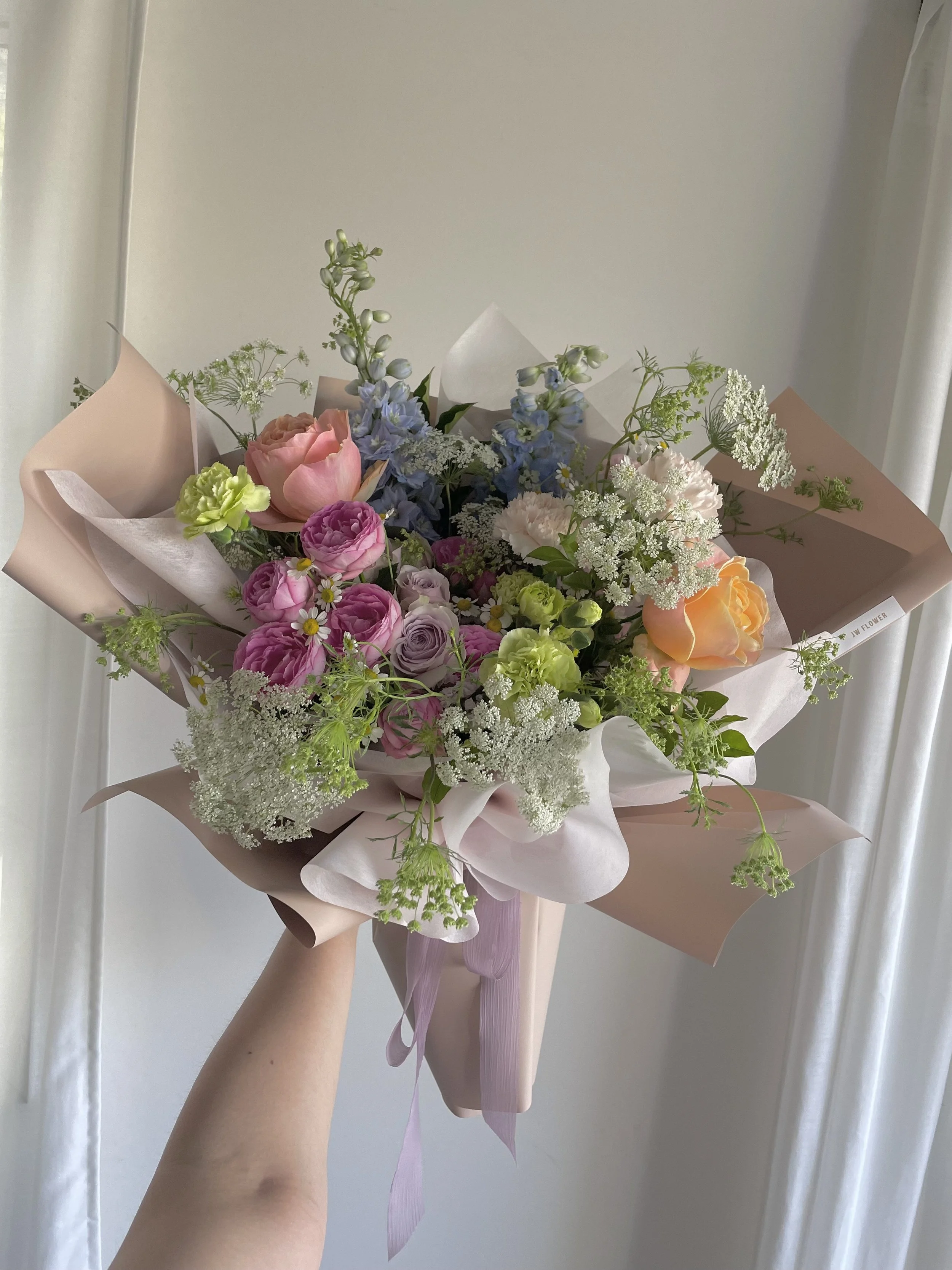 Hand holding a bouquet of assorted pastel-colored flowers, including pink roses, purple lisianthus, white baby’s breath, and green foliage, wrapped in pink and beige paper with a pink ribbon.