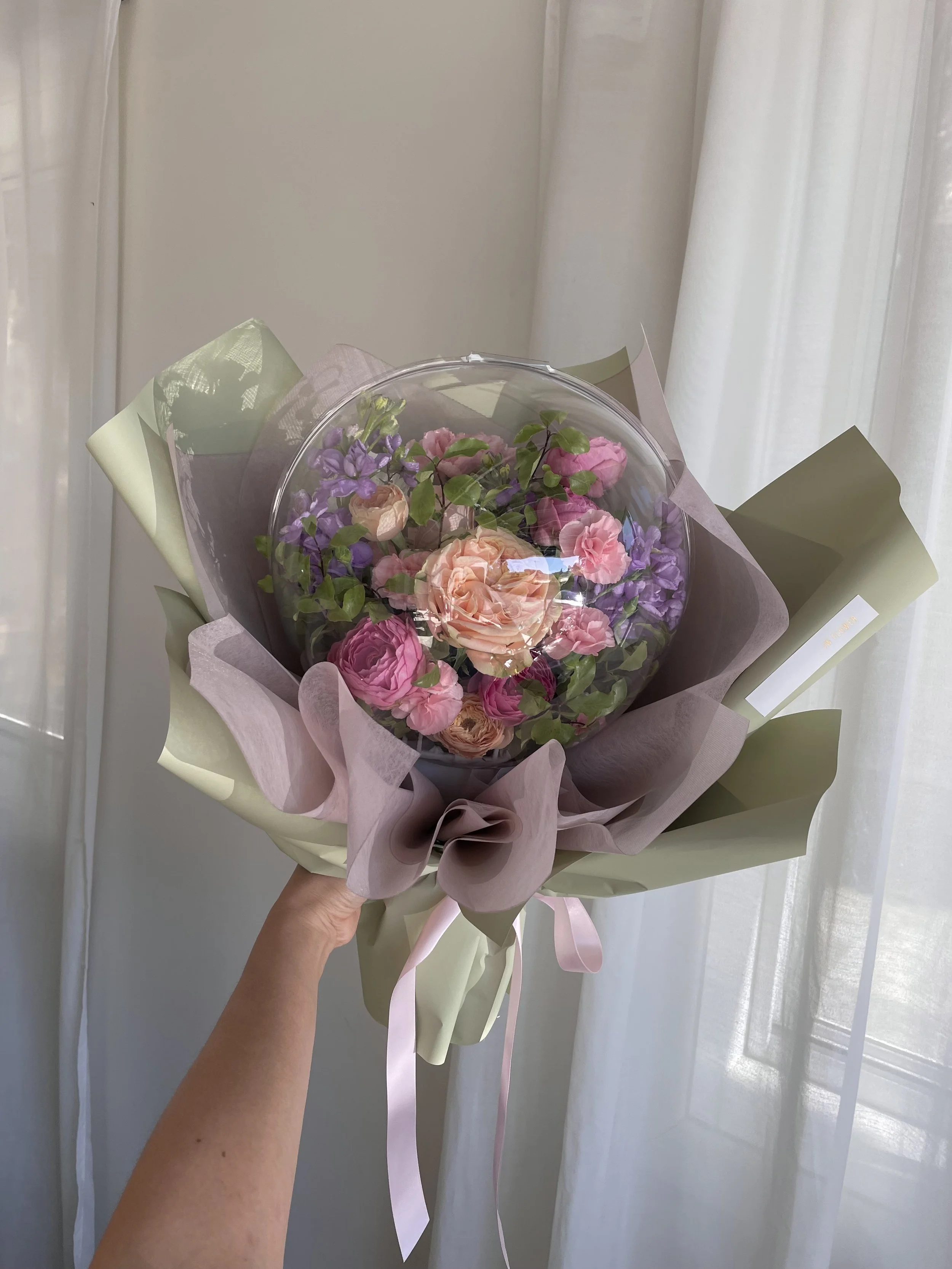 Hand holding a bouquet of pink and purple flowers wrapped in pastel tissue paper and green wrapping paper, with a domed glass cover on top.