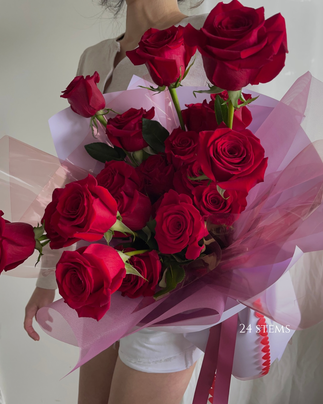 A person holding a large bouquet of red roses wrapped in pink and purple translucent paper.