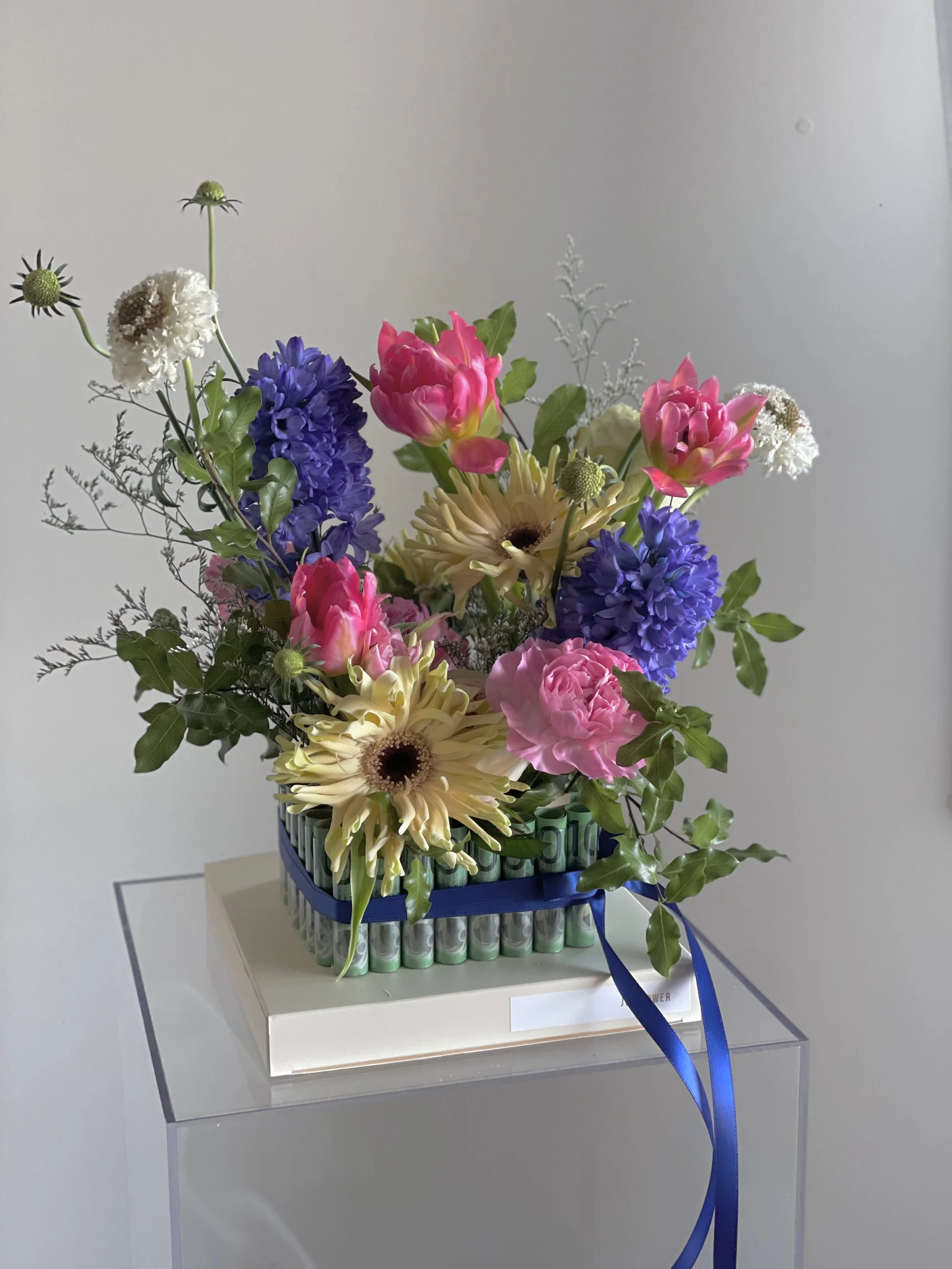 Colorful floral arrangement with pink, purple, white, and yellow flowers in a basket, wrapped with a blue ribbon, placed on a white box.