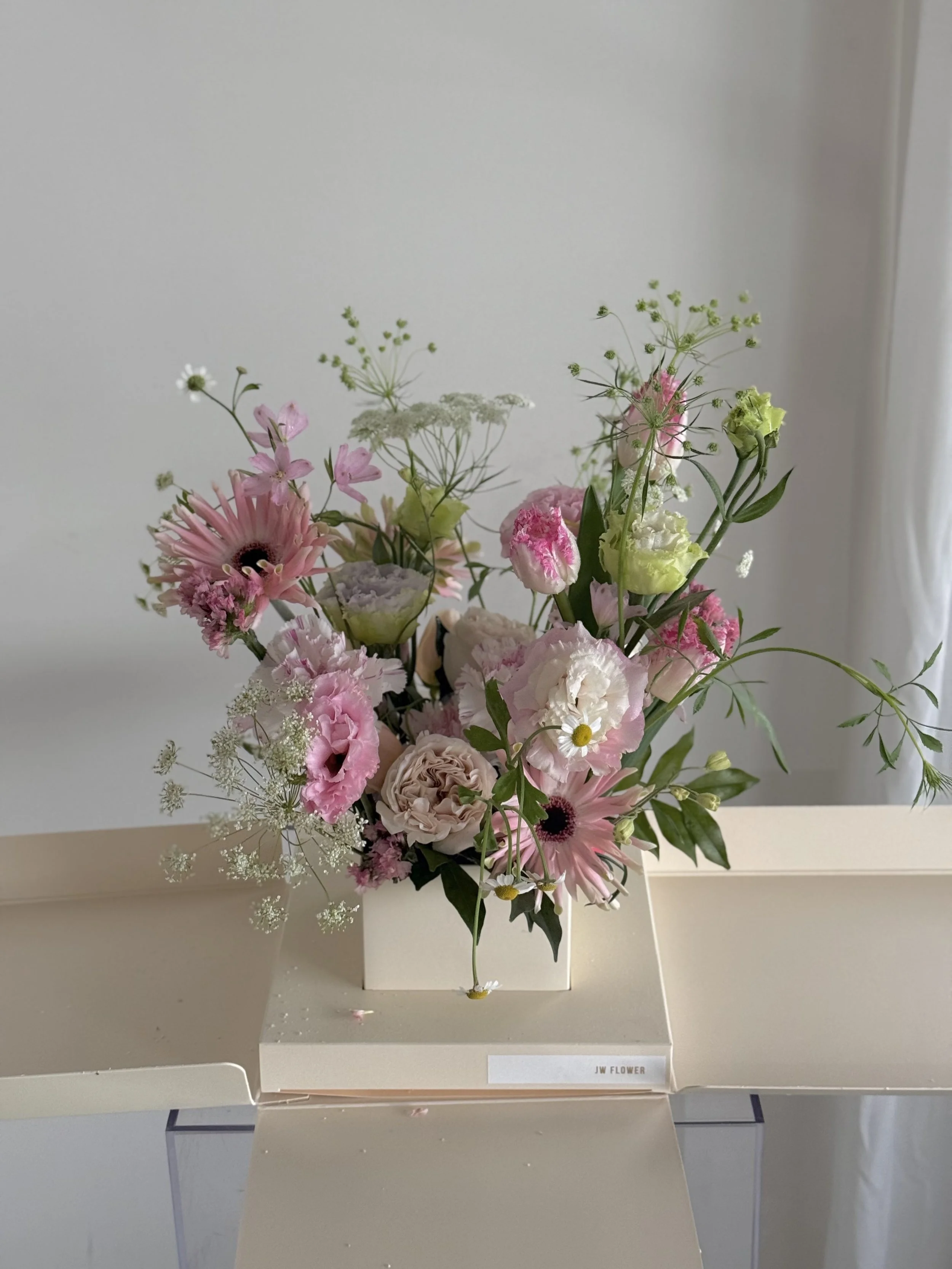 Pink and white mixed floral arrangement in a beige square vase, placed on a white box, on a light-colored surface in front of a light-colored wall with a curtain on the right.