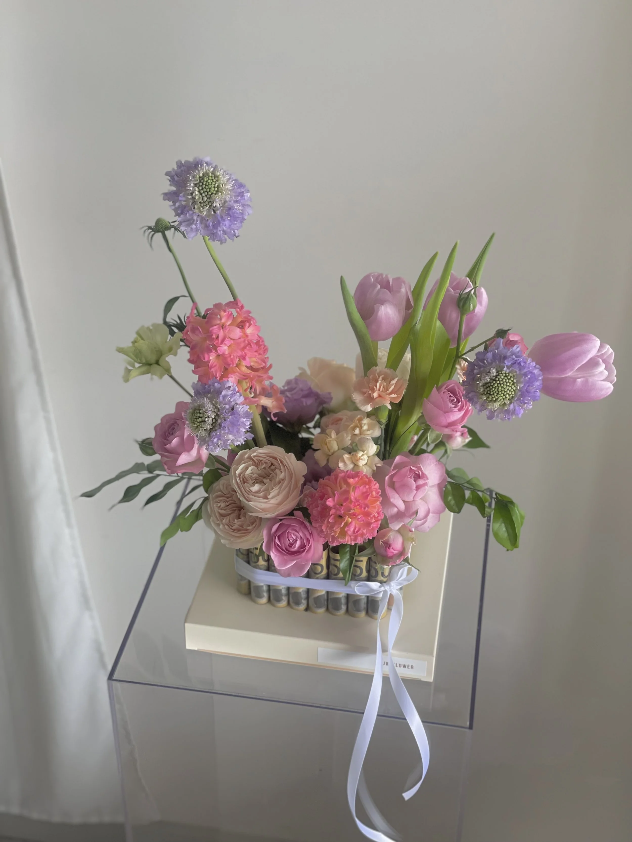 Colorful flower arrangement with pink tulips, roses, and purple scabiosa in a clear vase, displayed on a white box and pedestal.