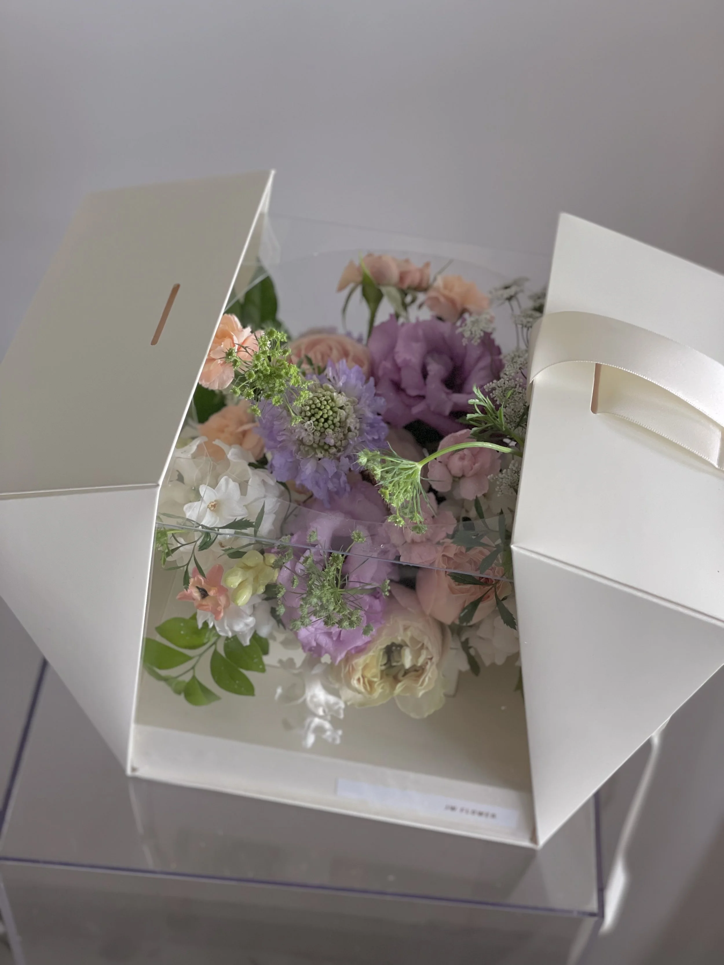 A floral arrangement with various pink, purple, white, and green flowers displayed in a clear box inside a white gift bag.