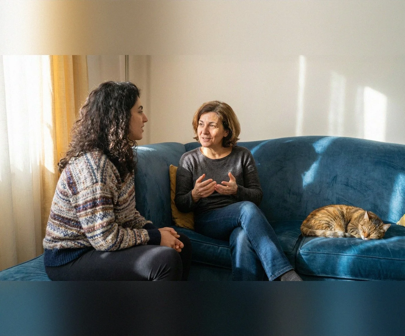 Two women sitting on a blue couch, engaged in conversation; one woman has dark, curly hair and the other has short, light brown hair. A sleeping tabby cat is on the couch beside them.