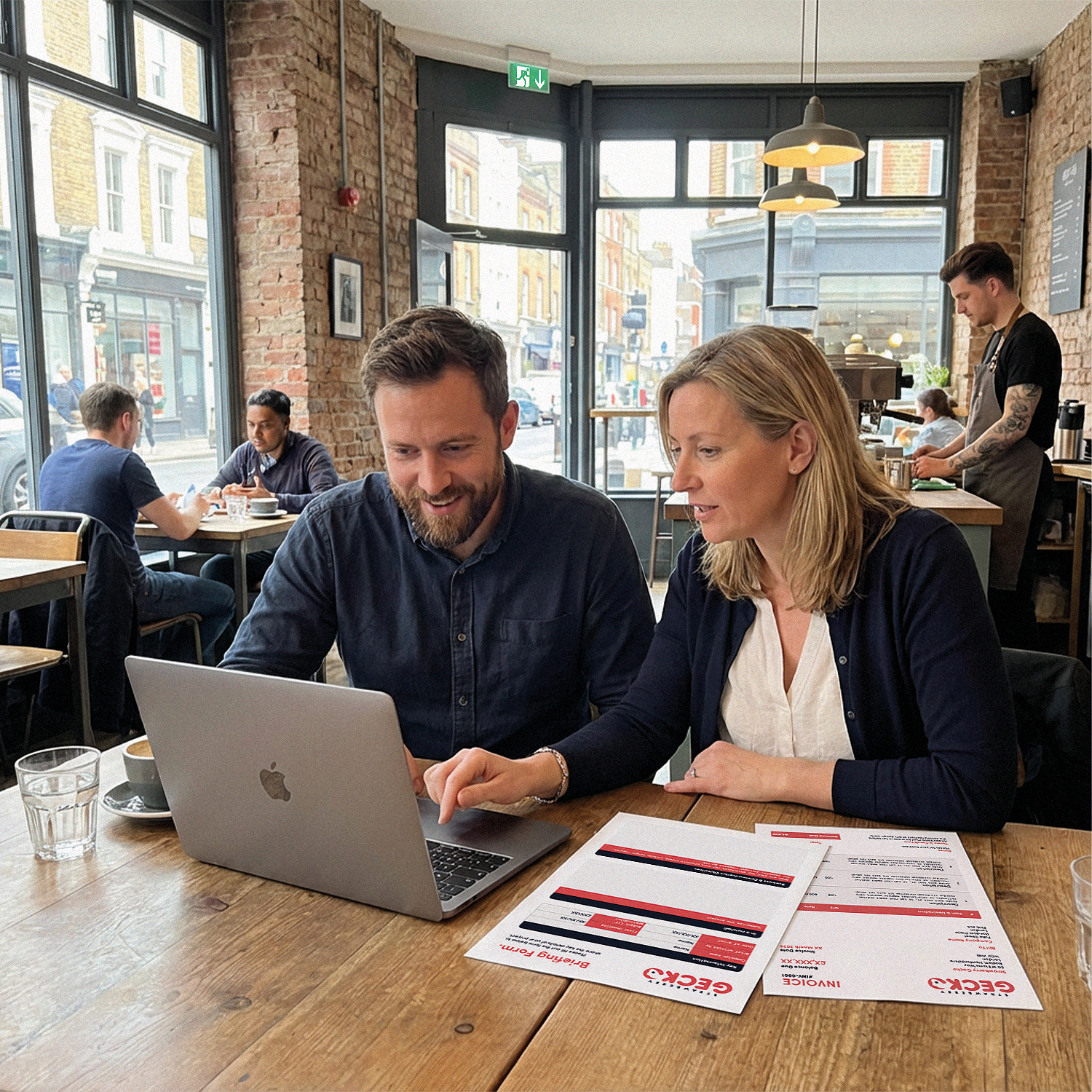 A man and a woman sitting at a wooden table in a cozy coffee shop, looking at a laptop together. There are menus on the table, and the background shows other customers, brick walls, large windows, and a barista working behind the counter.