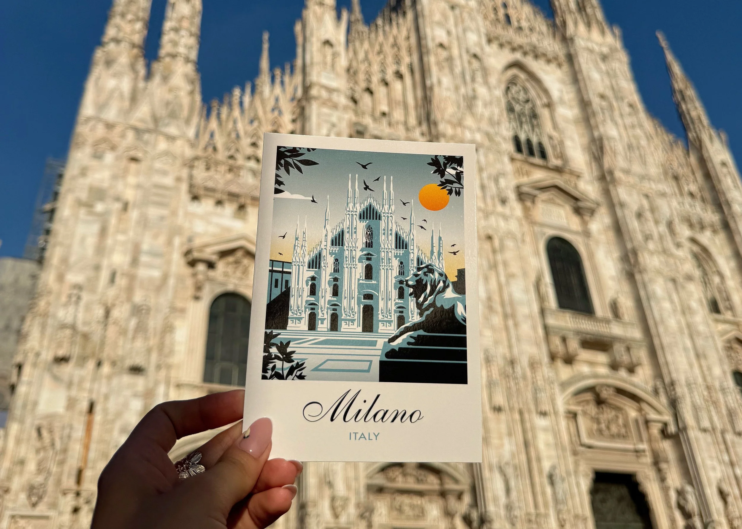 A person holding a postcard with an illustration of Milan Cathedral, with the word "Milano" and "Italy" written below, in front of Milan Cathedral in Italy.