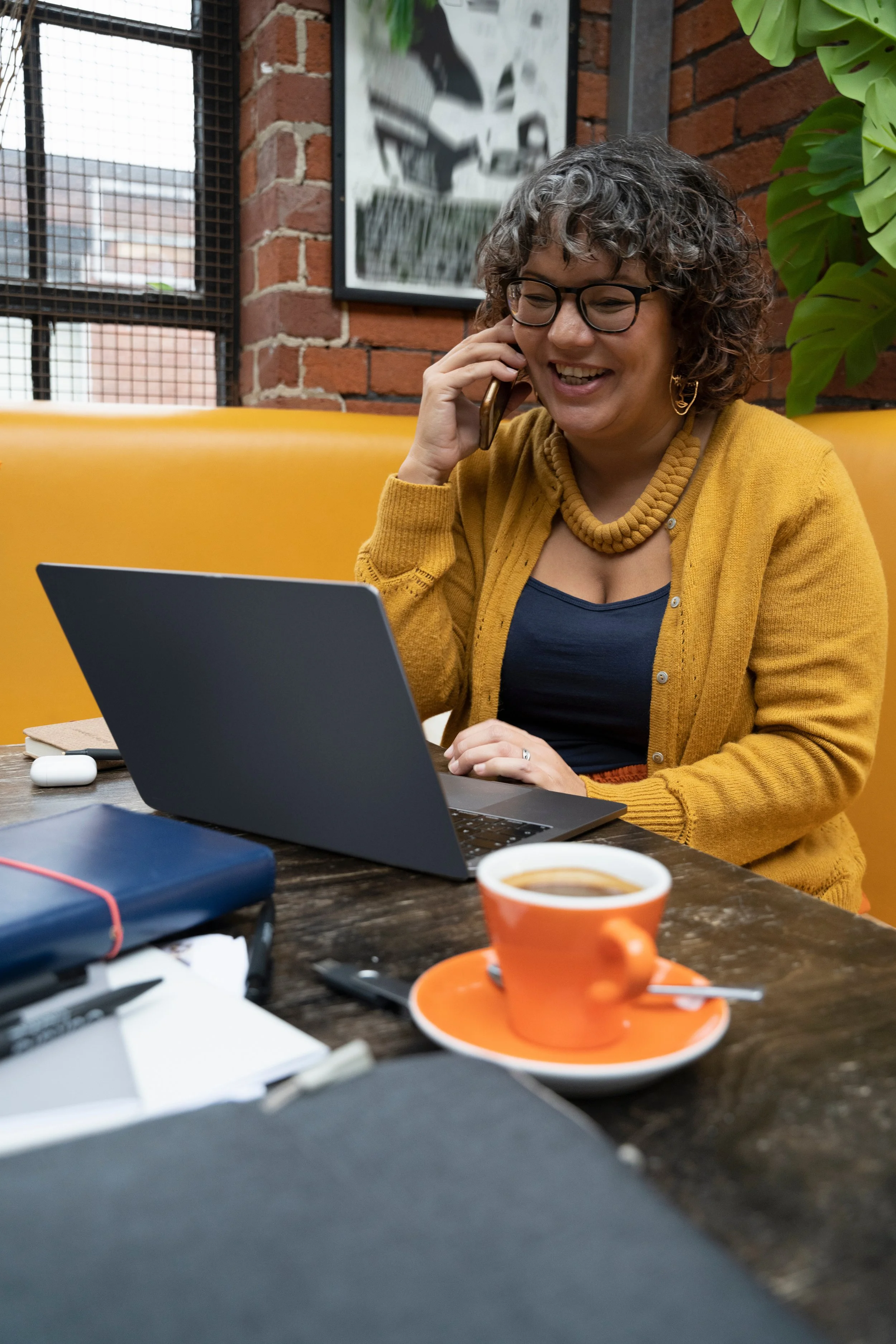 A woman with curly hair, glasses, and gold jewelry sitting at a wooden table in a cafe, talking on her phone, with a laptop, a cup of coffee on an orange saucer, and notebooks on the table.