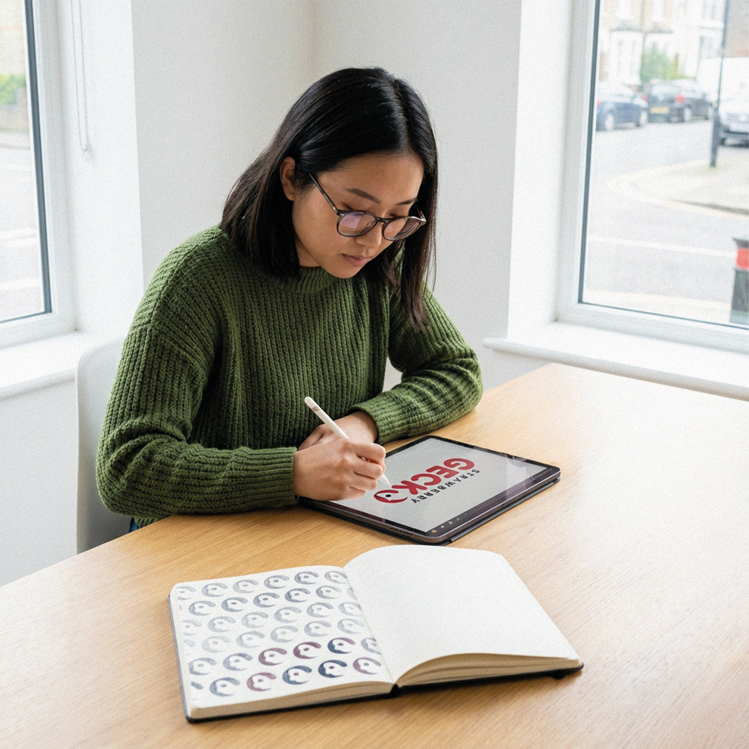 A woman with shoulder-length dark hair and glasses, wearing a green sweater, is sitting at a wooden table writing on a digital tablet that displays the style or logo of a brand called 'ECO' in large red letters. An open sketchbook with various logo designs is also on the table, and she is using a stylus. There are large windows behind her showing a street scene with parked cars.