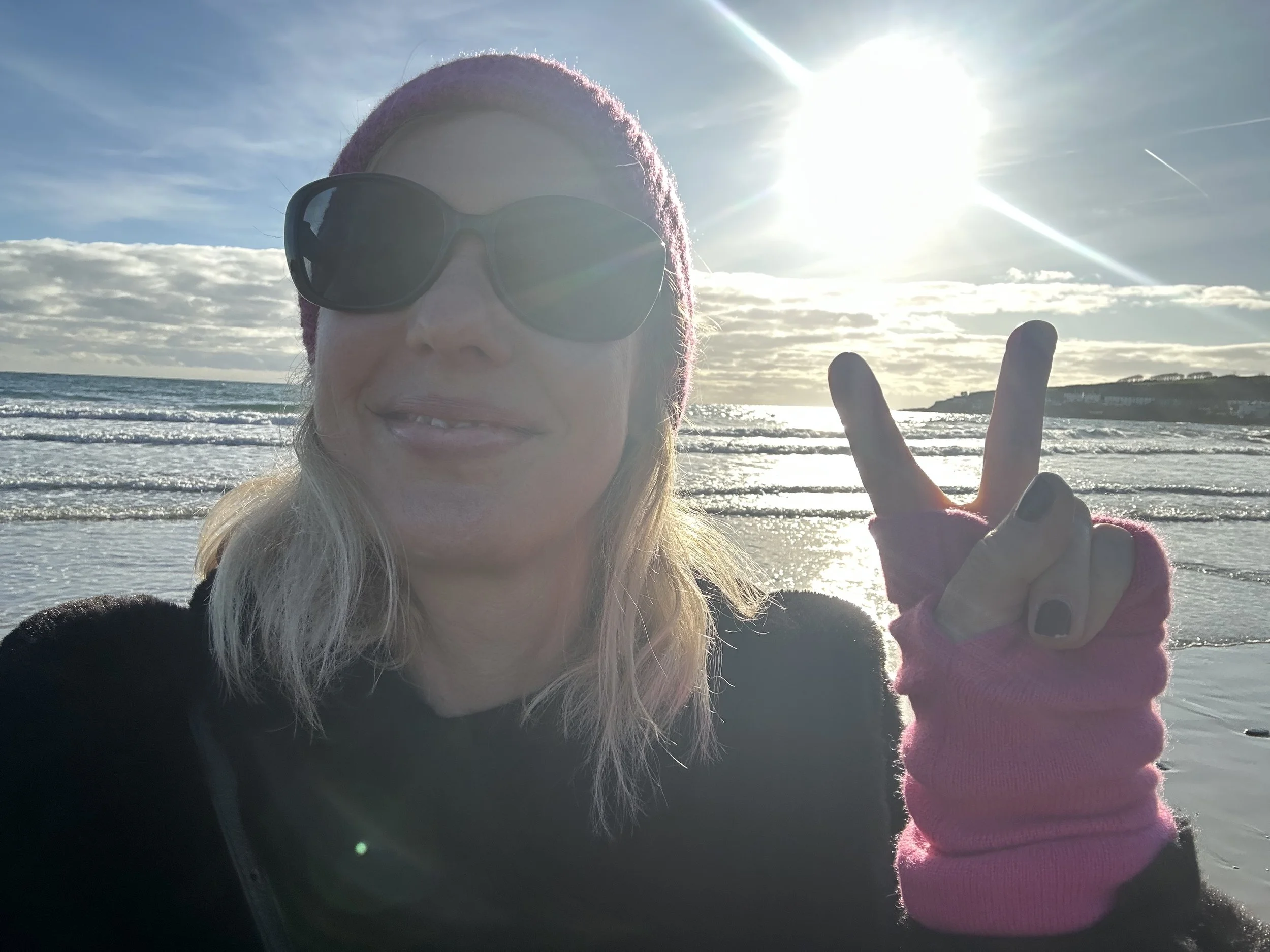 Woman wearing sunglasses and a pink beanie taking a selfie on the beach with the ocean and sky in the background, making a peace sign with her hand.