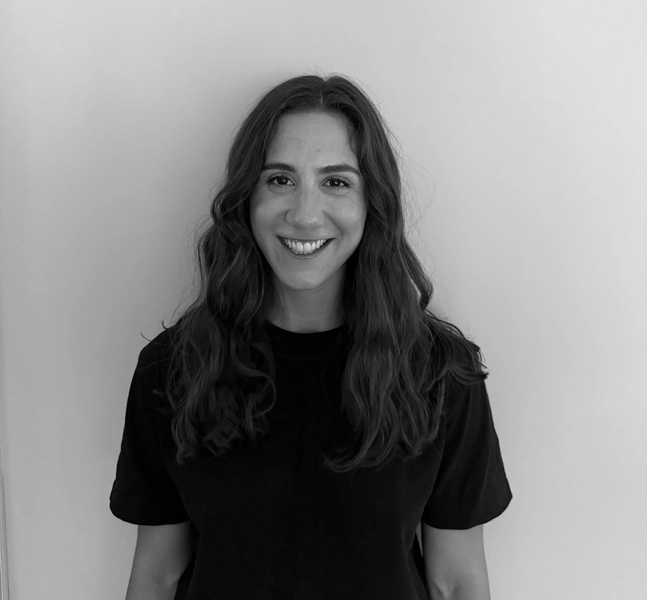 Black and white photo of Angelique, a woman with long wavy hair, smiling, wearing a black t-shirt, against a plain wall.