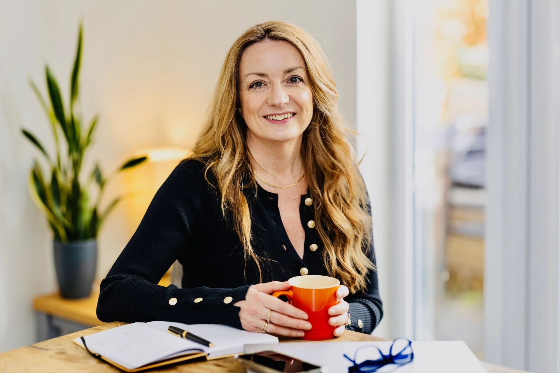 A woman with long red hair smiling and holding an orange coffee mug while sitting at a wooden table with an open notebook and a pair of glasses.