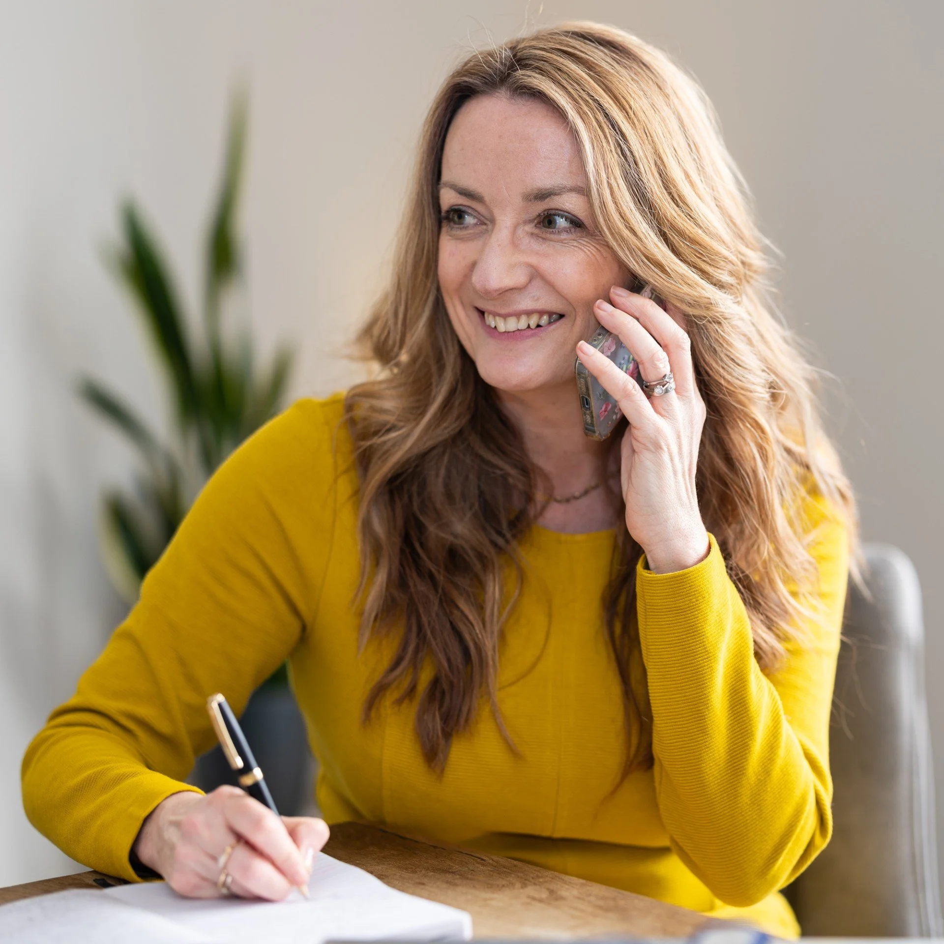 A woman with long, wavy reddish-brown hair wearing a yellow long-sleeved shirt, smiling while talking on a mobile phone, sitting at a desk with a pen and paper, in a bright indoor setting.