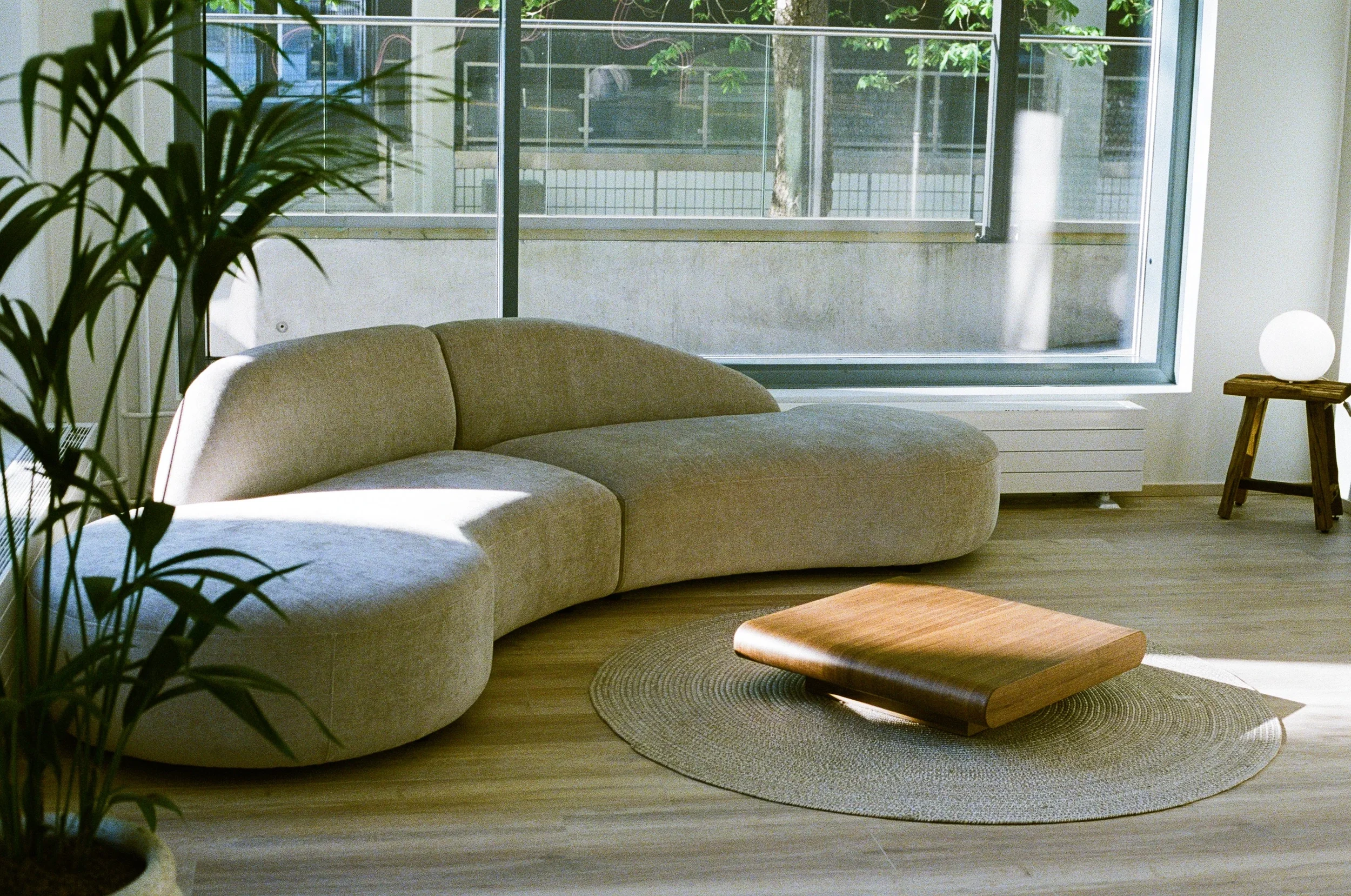Moderni olohuone suuri ikkunan edessä, beige kaareva sohva, pyöreä matto ja pieni puinen pöytä, jossa on lamppu, luonnonvalossa.
