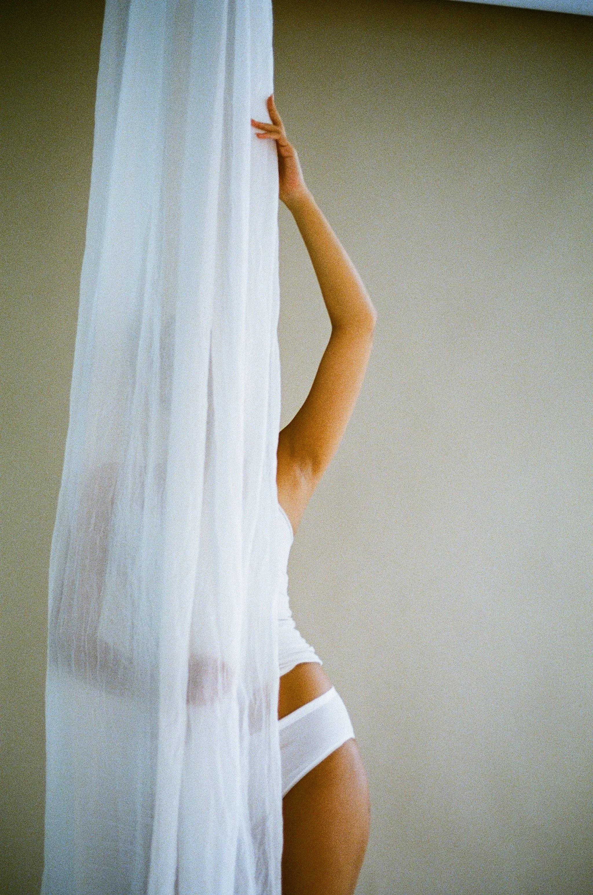 Naista osittain peitetynä valkoisen verhotarvittoman verhon takana, keskellä keho ja yksi käsi ulottuu ylös.