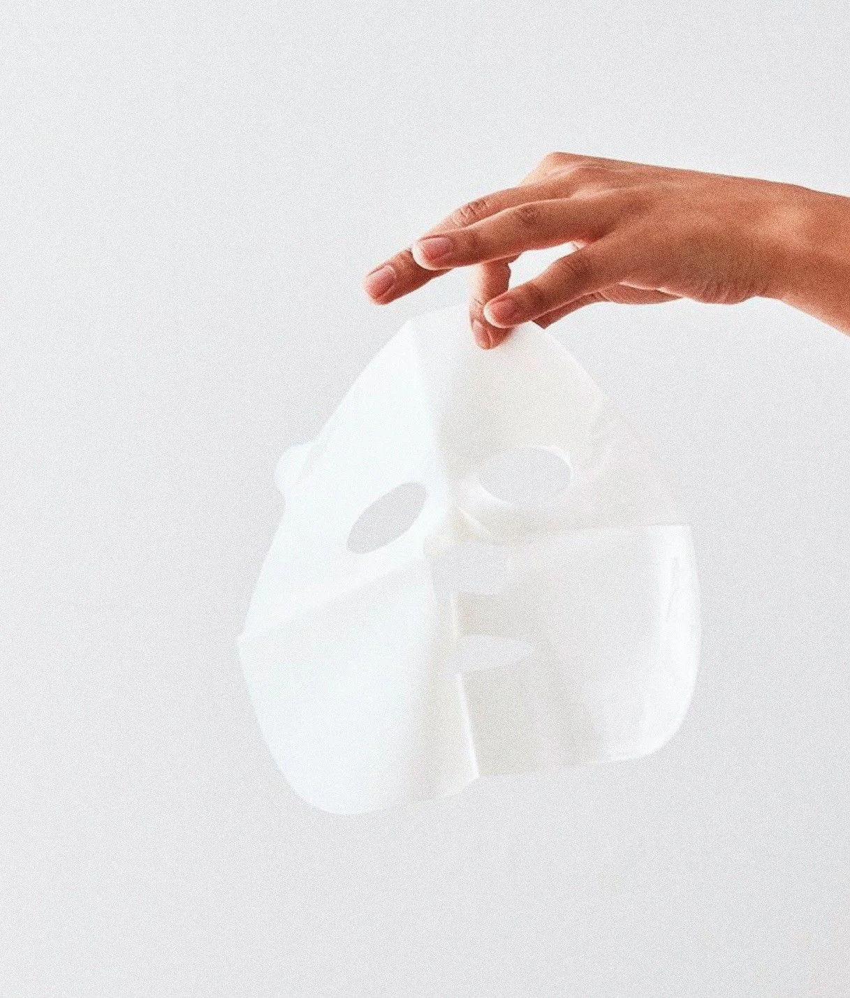 Käsin pidetään valkoinen kasvomaski, taustalla valkoinen seinä.