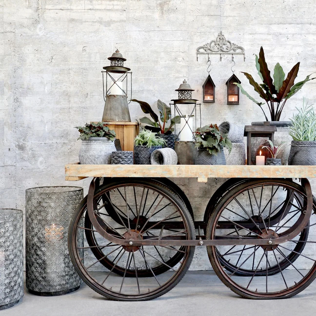 Wetterfahne auf einem alten Holzkorb, mit Tipi-Lichtern, Pflanzen in Töpfen und Laternen, vor einer weißen Wand.