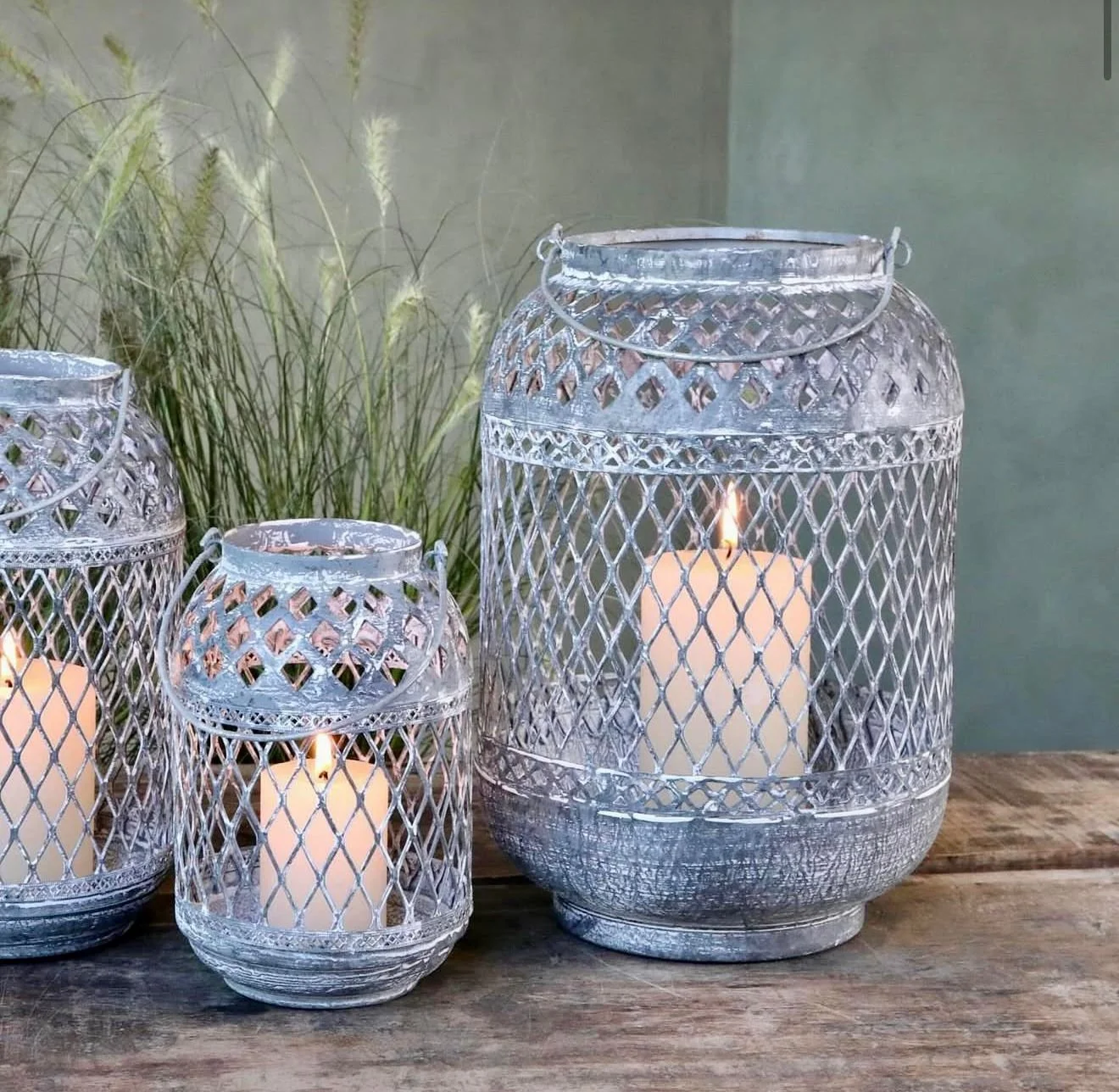 Drei silberne Laternen mit weißen Kerzen, die flackern, auf einem Holztisch, mit Pflanzen im Hintergrund.