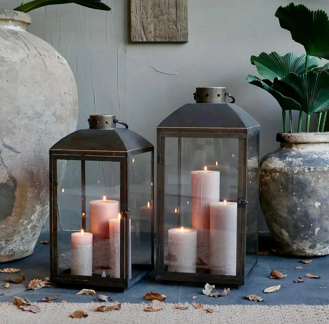 Zwei große Metall-Kerzenlaternen mit brennenden Kerzen, umgeben von großen antiken Tontöpfen mit grünen Pflanzen und verstreuten Herbstlaub auf dem Boden.