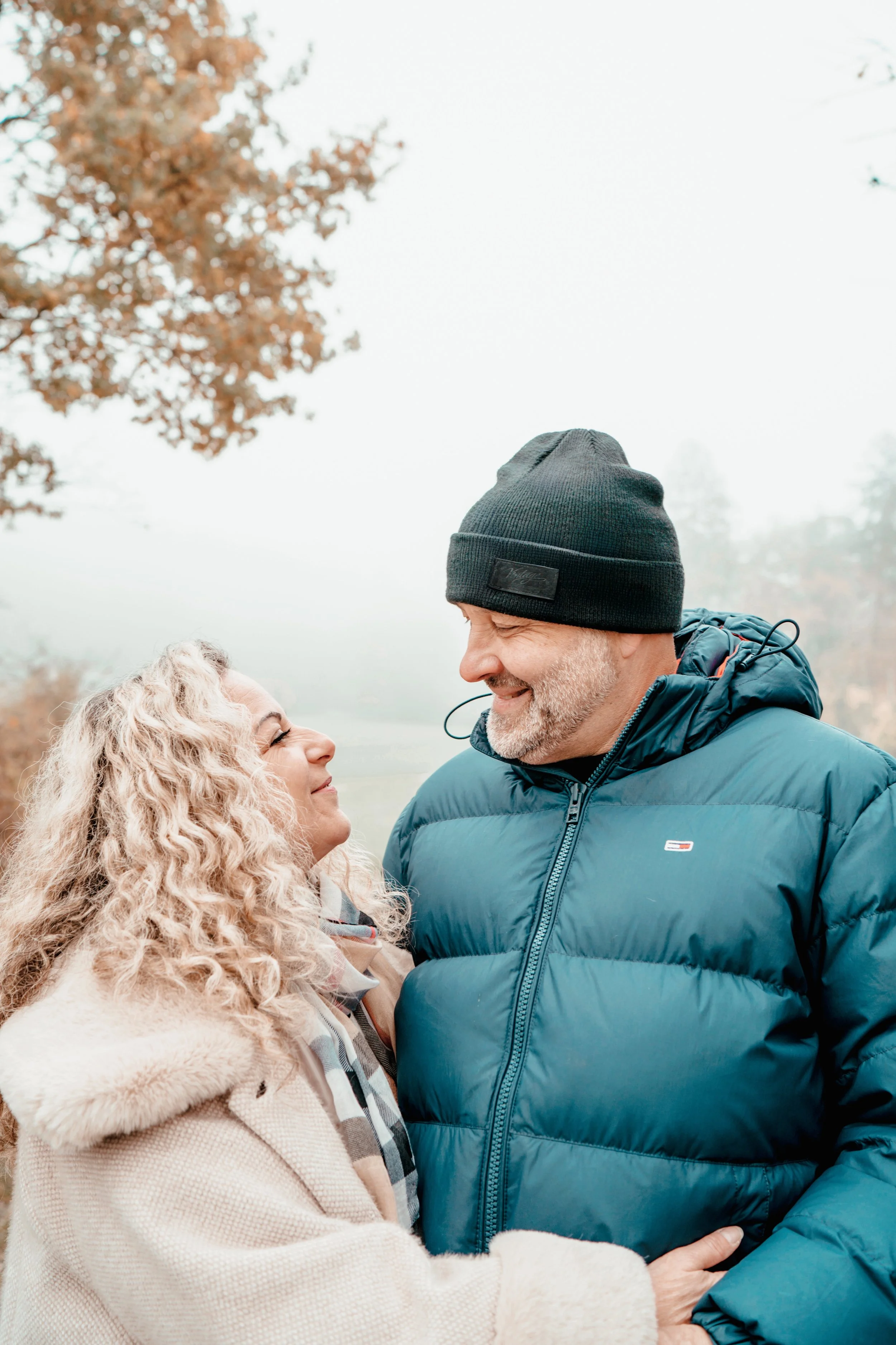 Ein lachendes älteres Paar, das sich im Freien umarmt, bei kühlem Wetter, mit bewölktem Himmel, Bäumen und Blättern im Herbst.