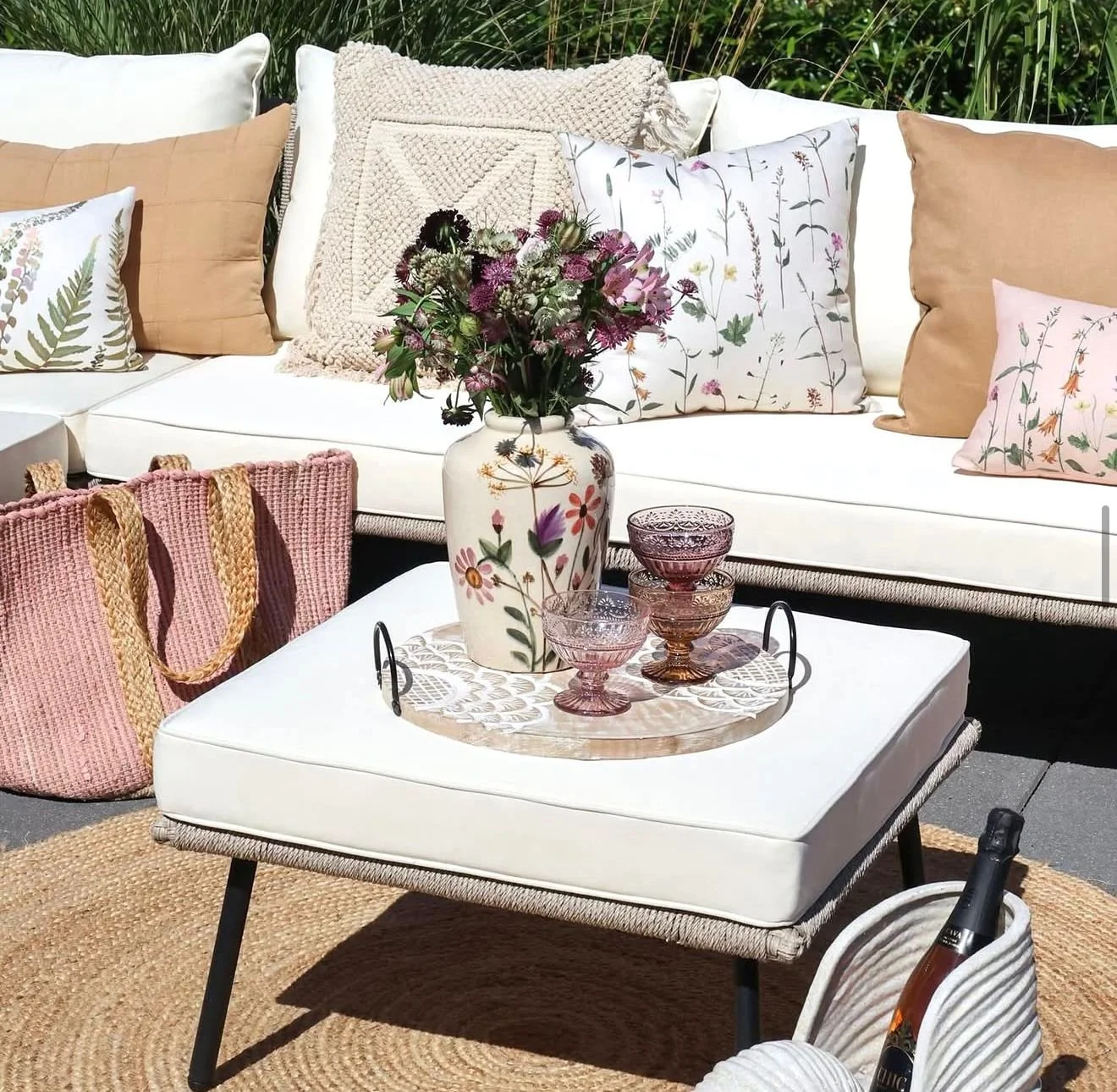 Gemütliche Sitzecke im Außenbereich mit weißen Kissen und bunten Kissen, ein Couchtisch mit Blumenvasen und Trinkgläsern, Umhängetasche und Flasche Sekt auf einem verzierten Tablett, Rattan-Untersetzer, weicher Teppichboden.