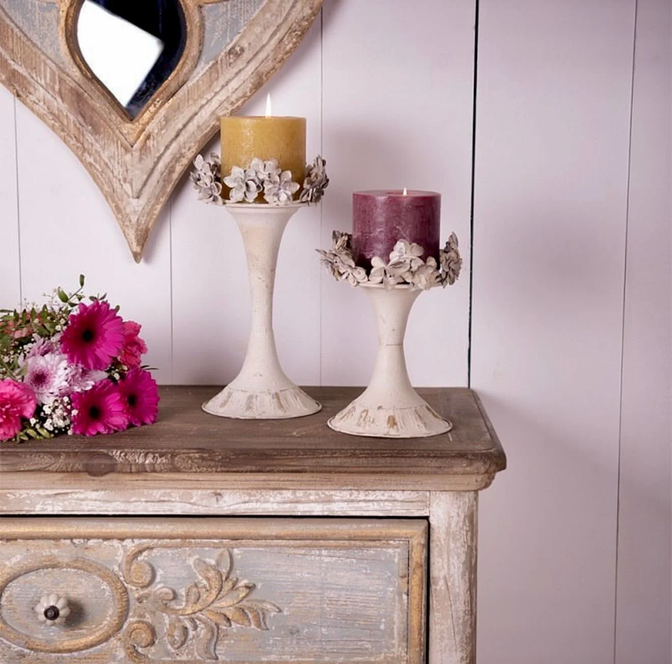 Zwei weiße Kerzen in dekorativen Kerzenständern auf einer Holzkommode, daneben ein Blumenstrauß mit pinken Blumen, im Hintergrund ein weißes Wandpaneel.