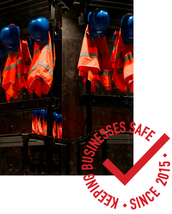 Blue hard hats and orange safety vests hang on a dark rack. A red emblem reads "Keeping Businesses Safe Since 2015," conveying safety and professionalism.