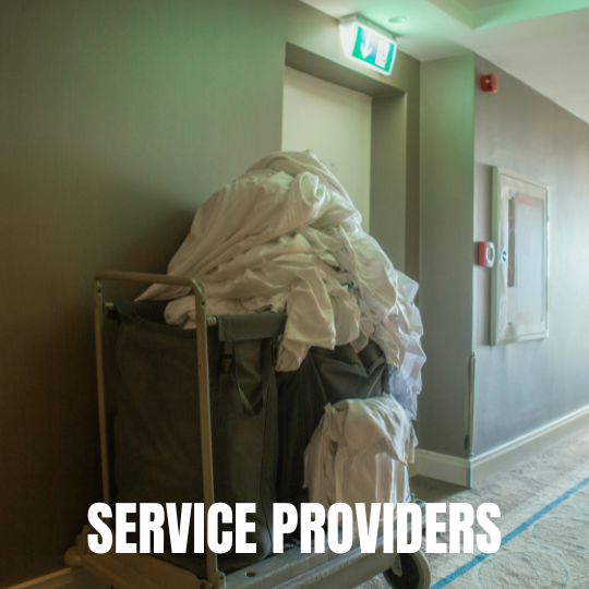 A cart filled with a large pile of white linens in a dimly lit hallway, under an exit sign. Text reads "Service Providers" at the bottom.
