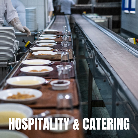 A catering conveyor belt with plates of food, utensils, and glasses in a busy kitchen setting. Text overlay reads 'Hospitality & Catering' in bold.