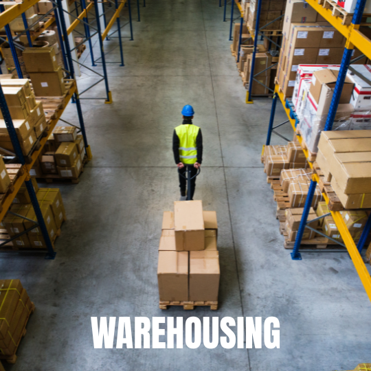 Aerial view of a warehouse with a worker in a blue helmet and yellow vest moving a pallet jack with boxes. Shelves are stacked with items. Text reads "Warehousing."
