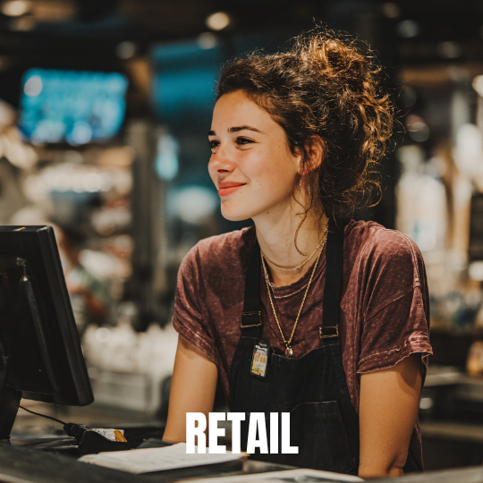 Smiling employee in a retail store, wearing a casual t-shirt and apron, stands at a checkout counter. The atmosphere is warm and inviting.