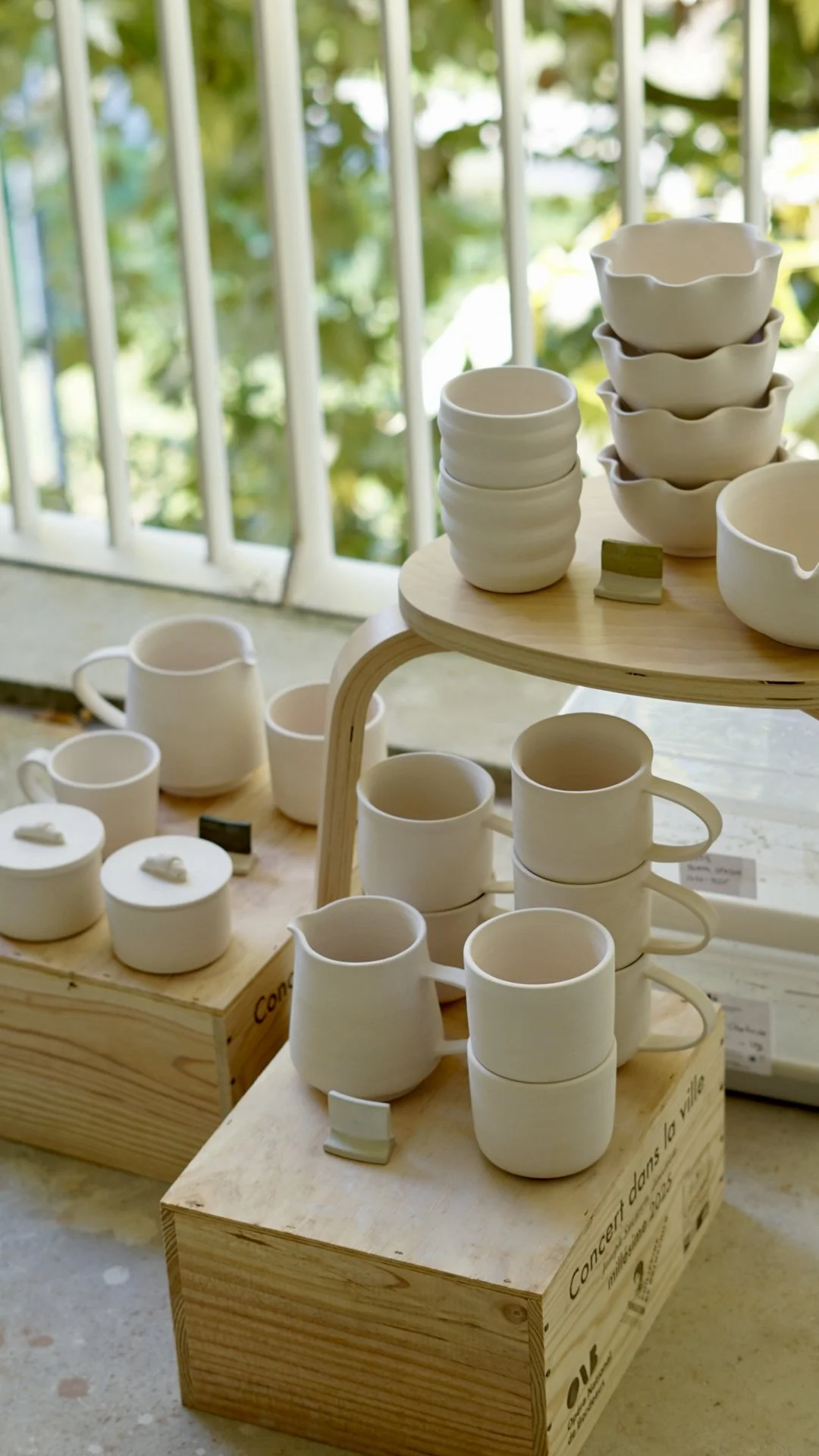 Stacked white ceramic bowls and mugs on wooden display stands outdoors against a green leafy background.