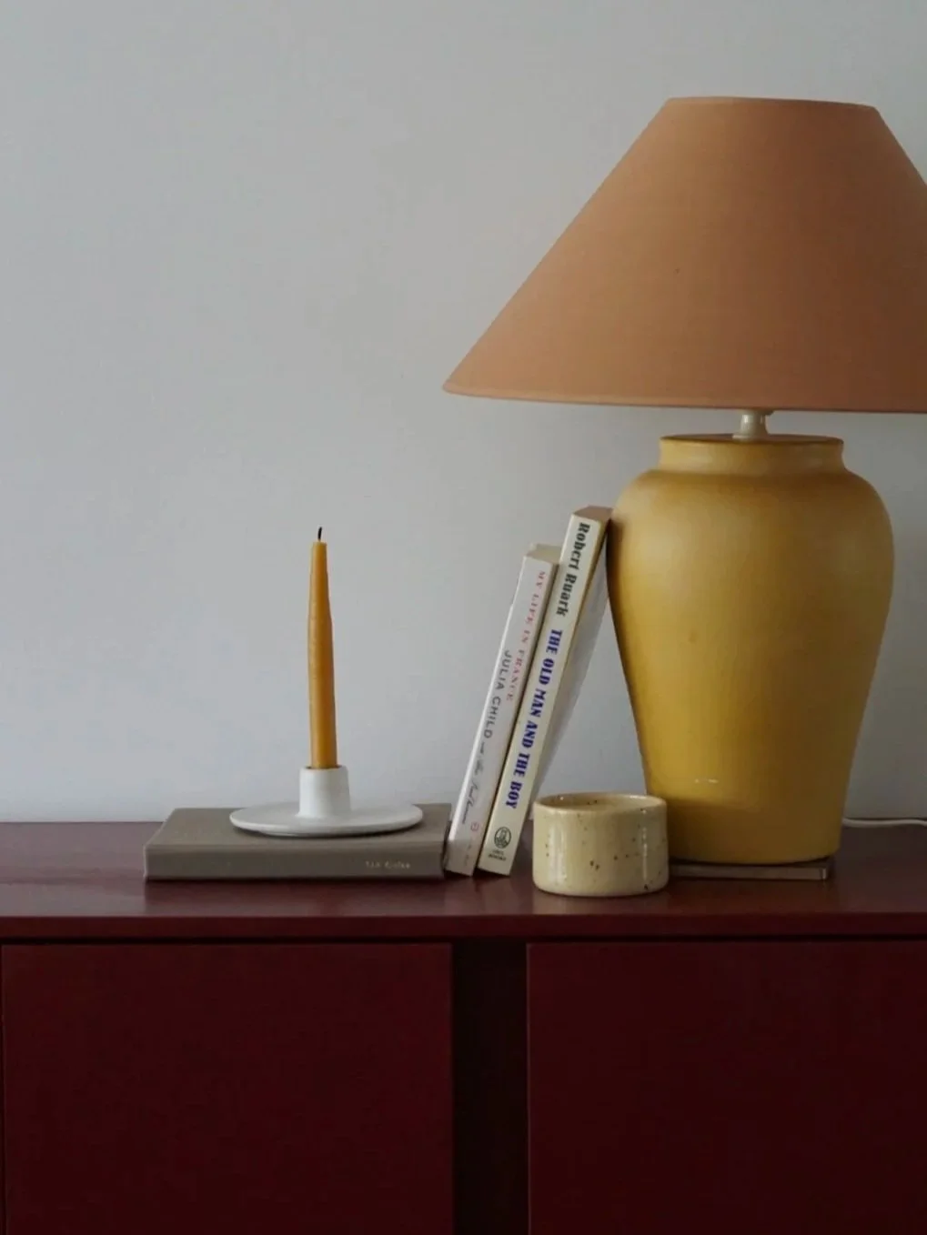 A wooden side table with a tall yellow candle on a white dish, a stack of three books leaning against a yellow ceramic lamp with a beige lampshade, and a small off-white ceramic cup in front of the lamp.