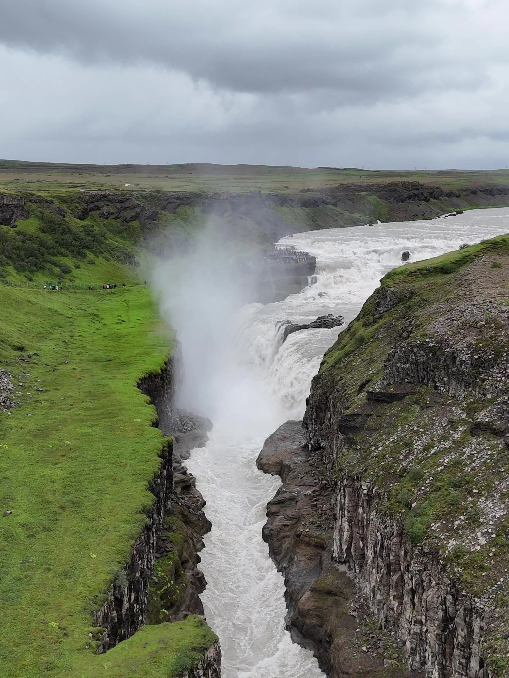 Icelandic waterfall with white water cascading through a deep rocky canyon, surrounded by green grassy terrain under a cloudy sky.