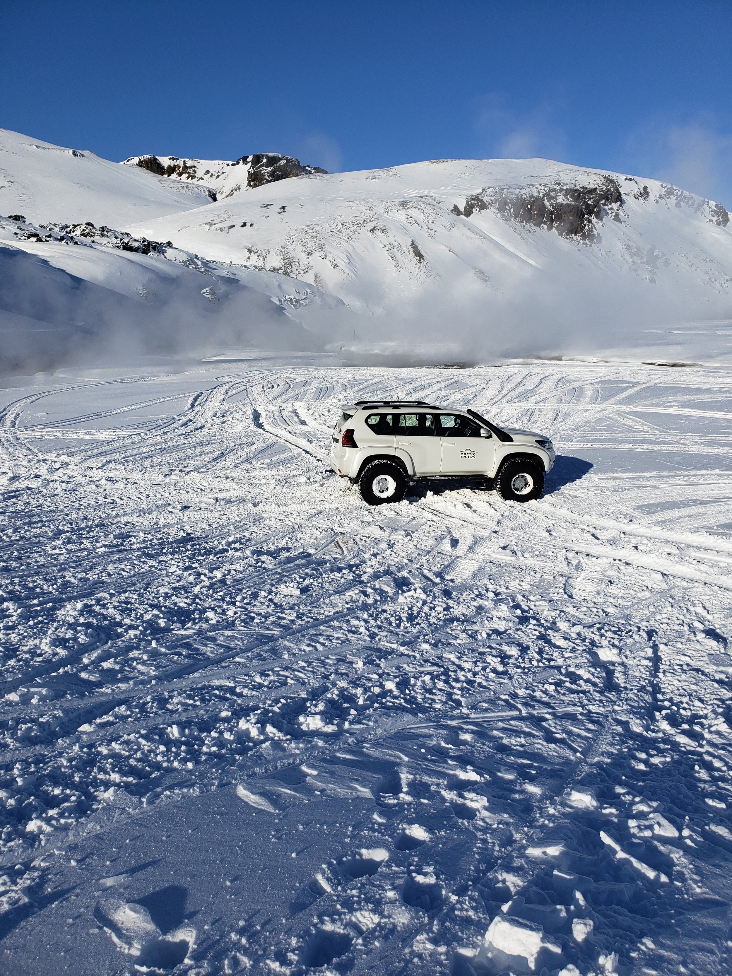 A white SUV parked on snow-covered terrain with tracks and footprints, with snow-covered mountains and a clear blue sky in the background.