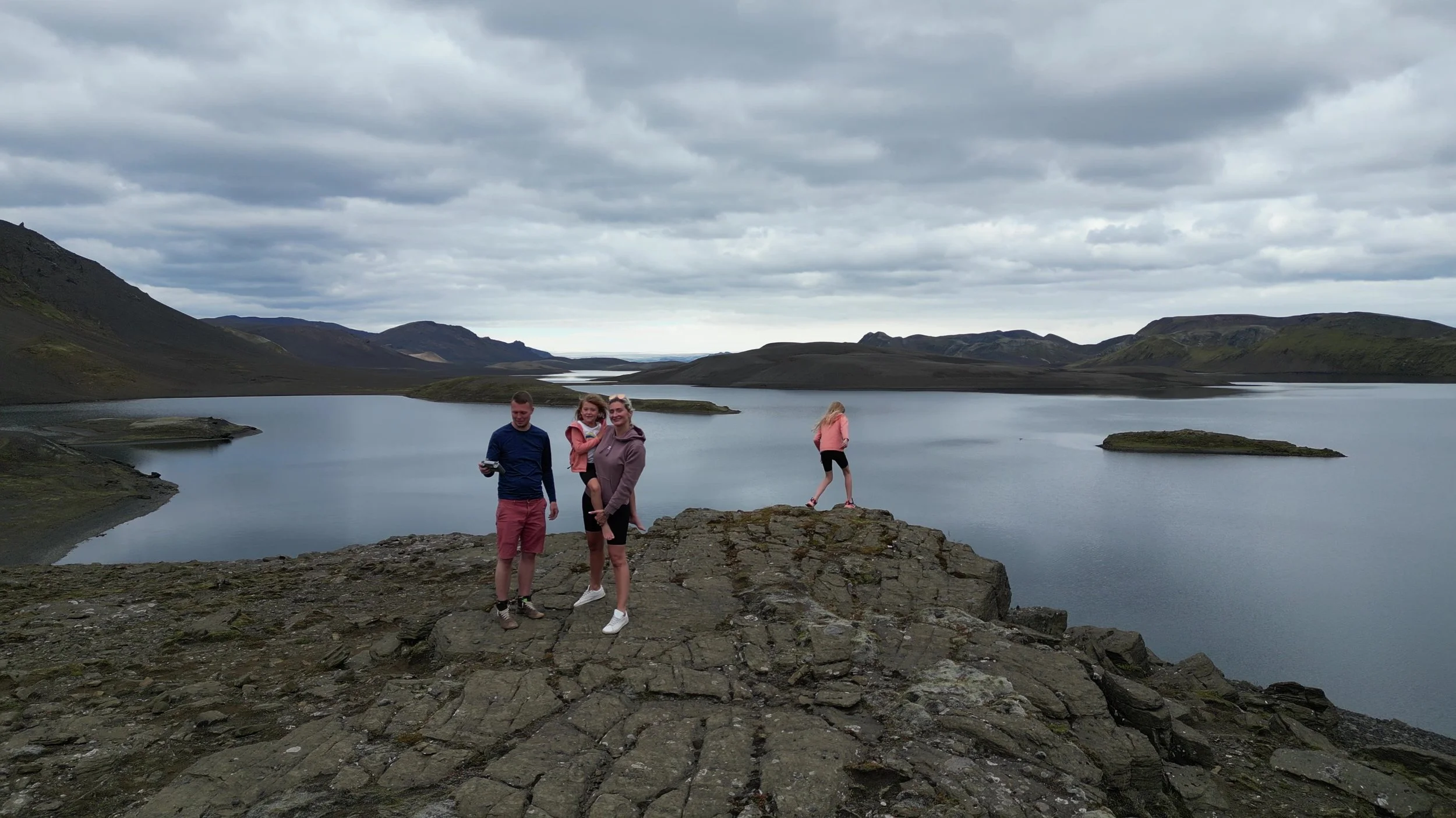 Four people standing and walking on a rocky outcrop overlooking a large lake with hills and cloudy sky in the background.