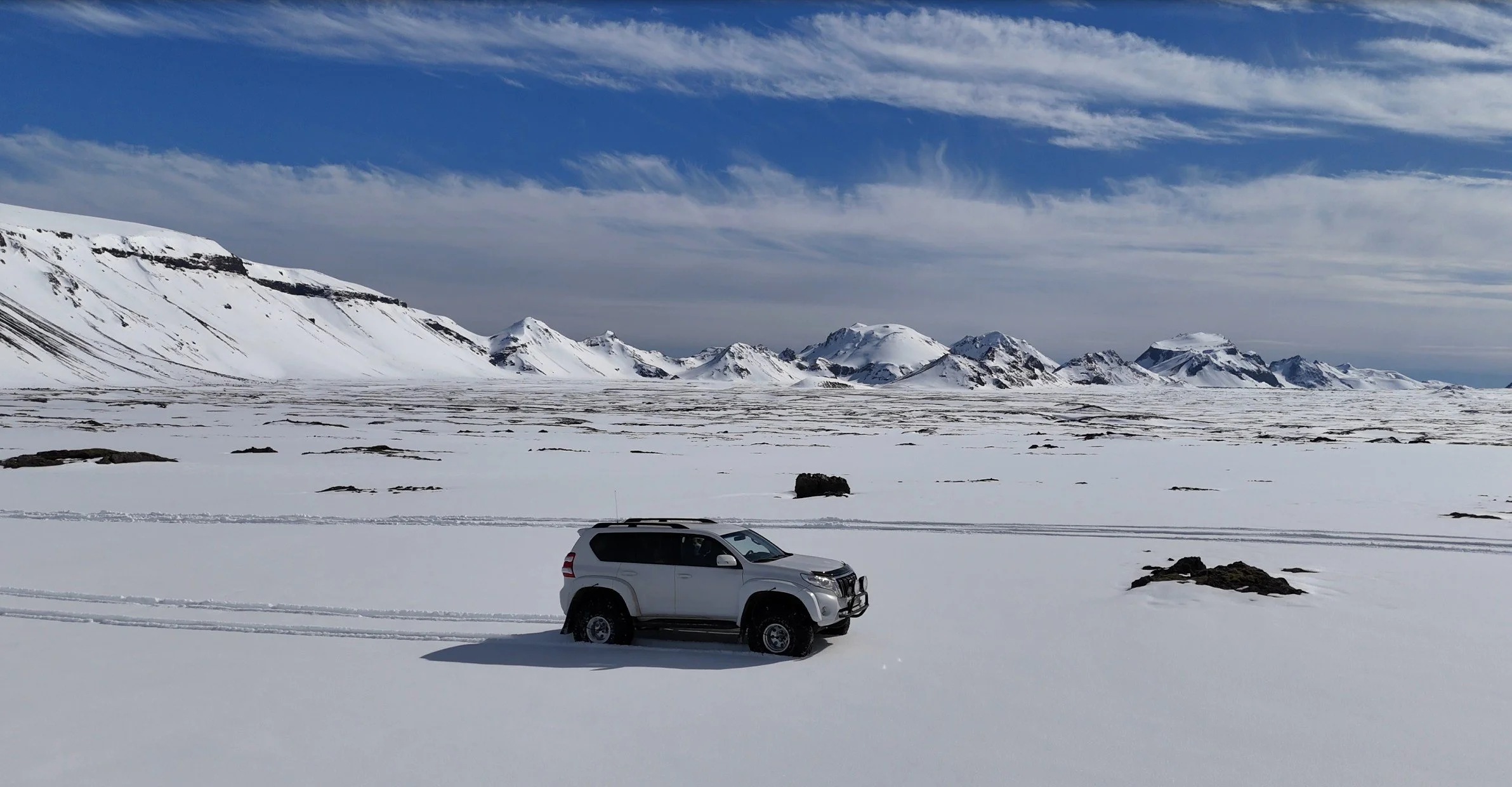 A white SUV parked on snow-covered terrain with snow-capped mountains in the background under a partly cloudy sky.