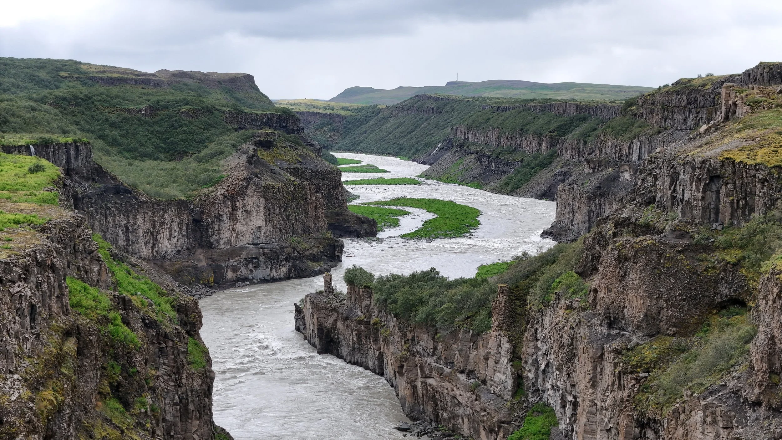 A river flowing through a deep, rugged canyon with rocky cliffs on either side, and distant green hills under a cloudy sky.