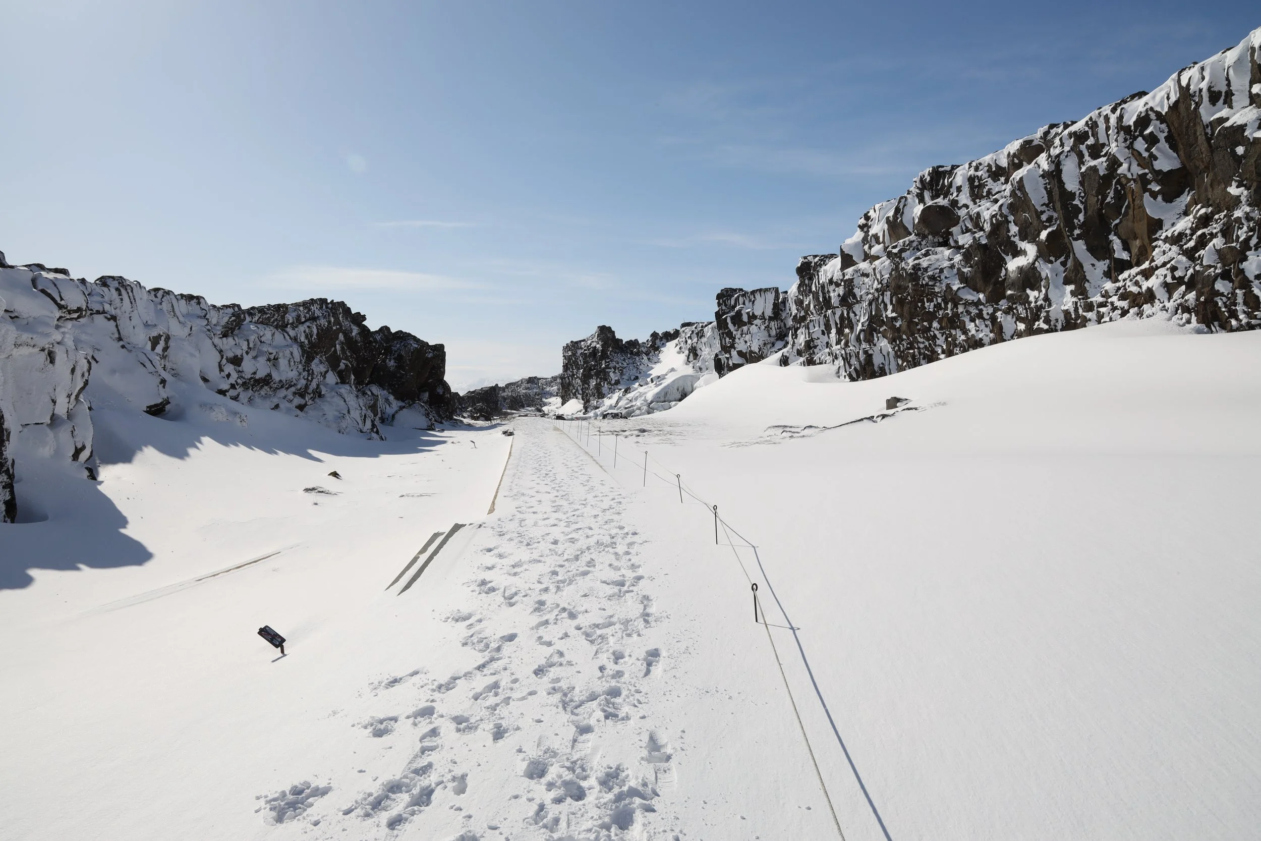 Snow-covered path flanked by rocky cliffs under a blue sky.