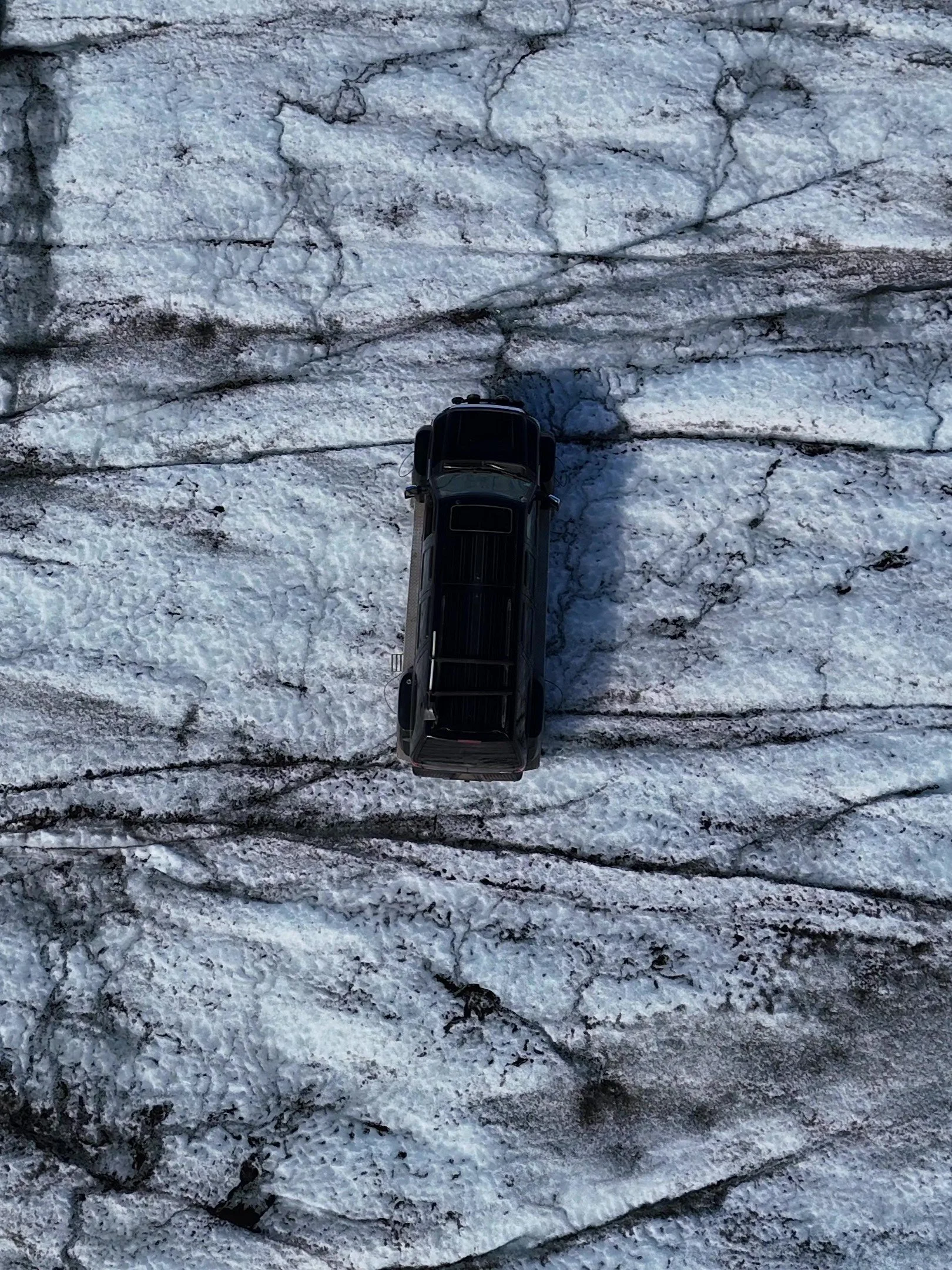 An overhead view of a black SUV parked on a snow-covered, uneven rocky surface.