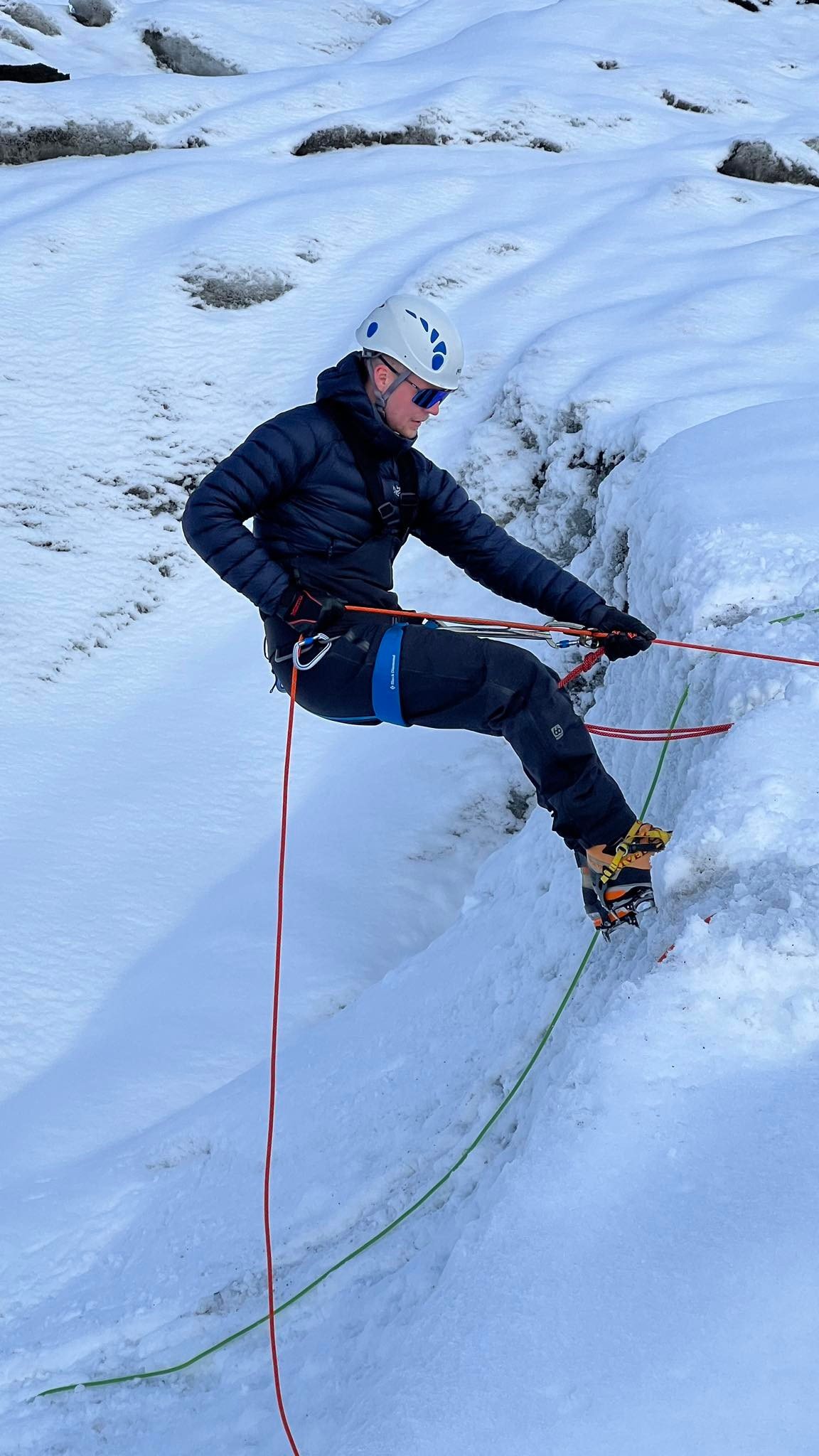 A man wearing a white helmet, blue sunglasses, and a dark winter outfit is ice climbing on a frozen wall, using climbing gear and ropes for safety.